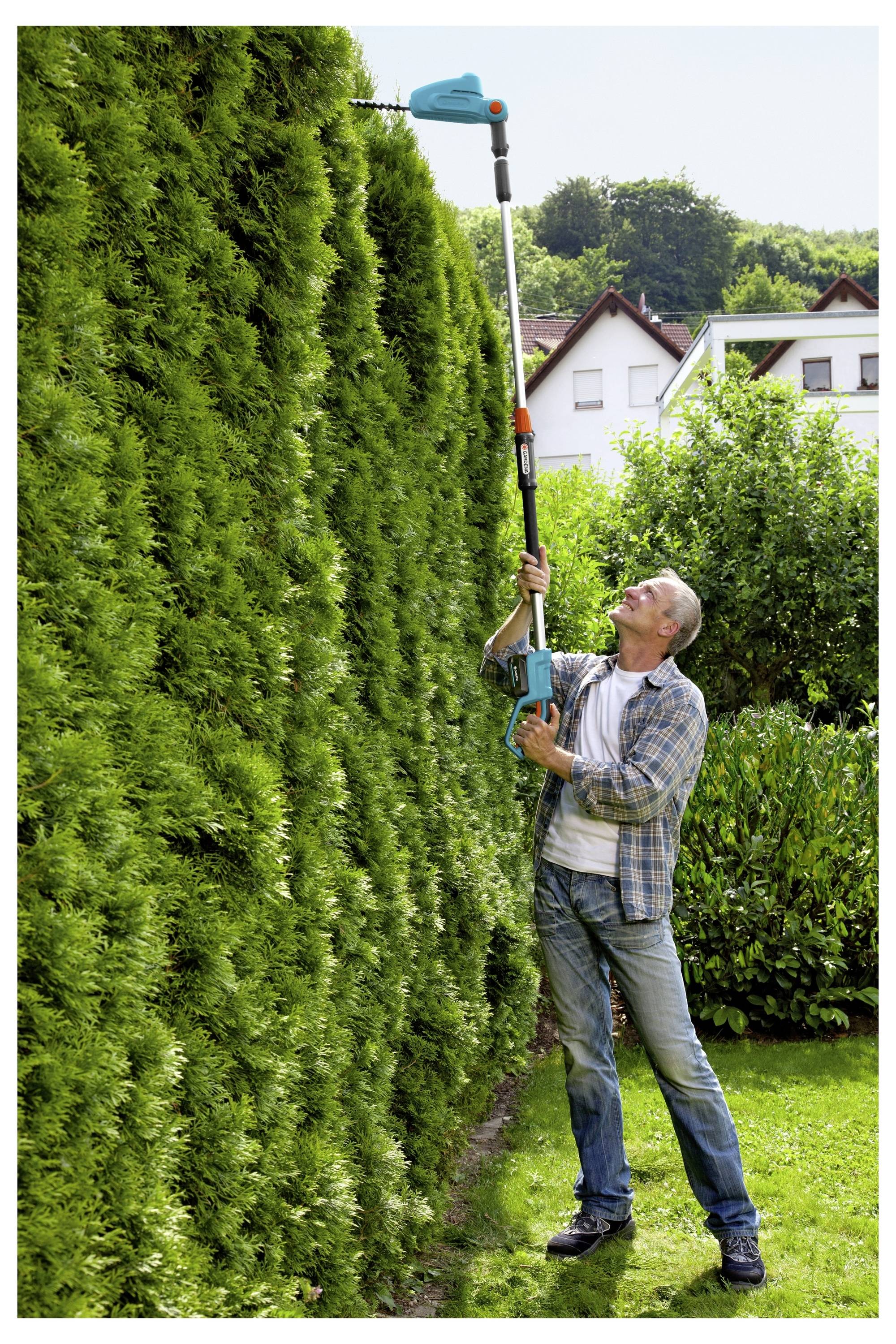 Mężczyzna przystrzyga wierzchołek wysokiego żywopłotu za pomocą elektrycznej przyciniarki z długim trzonkiem na terenie domu, otoczony drzewami i domami w tle.