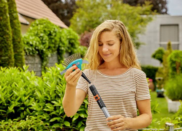 Kobieta w ogrodzie trzymająca małą grabie, otoczona bujną zielenią roślin, sugerująca czynności ogrodnicze.