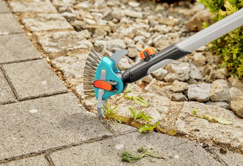 Narzędzie ogrodnicze usuwa chwasty między kostką brukową, prezentując zbliżenie jego zastosowania na ścieżce otoczonej kamieniami i zielenią.