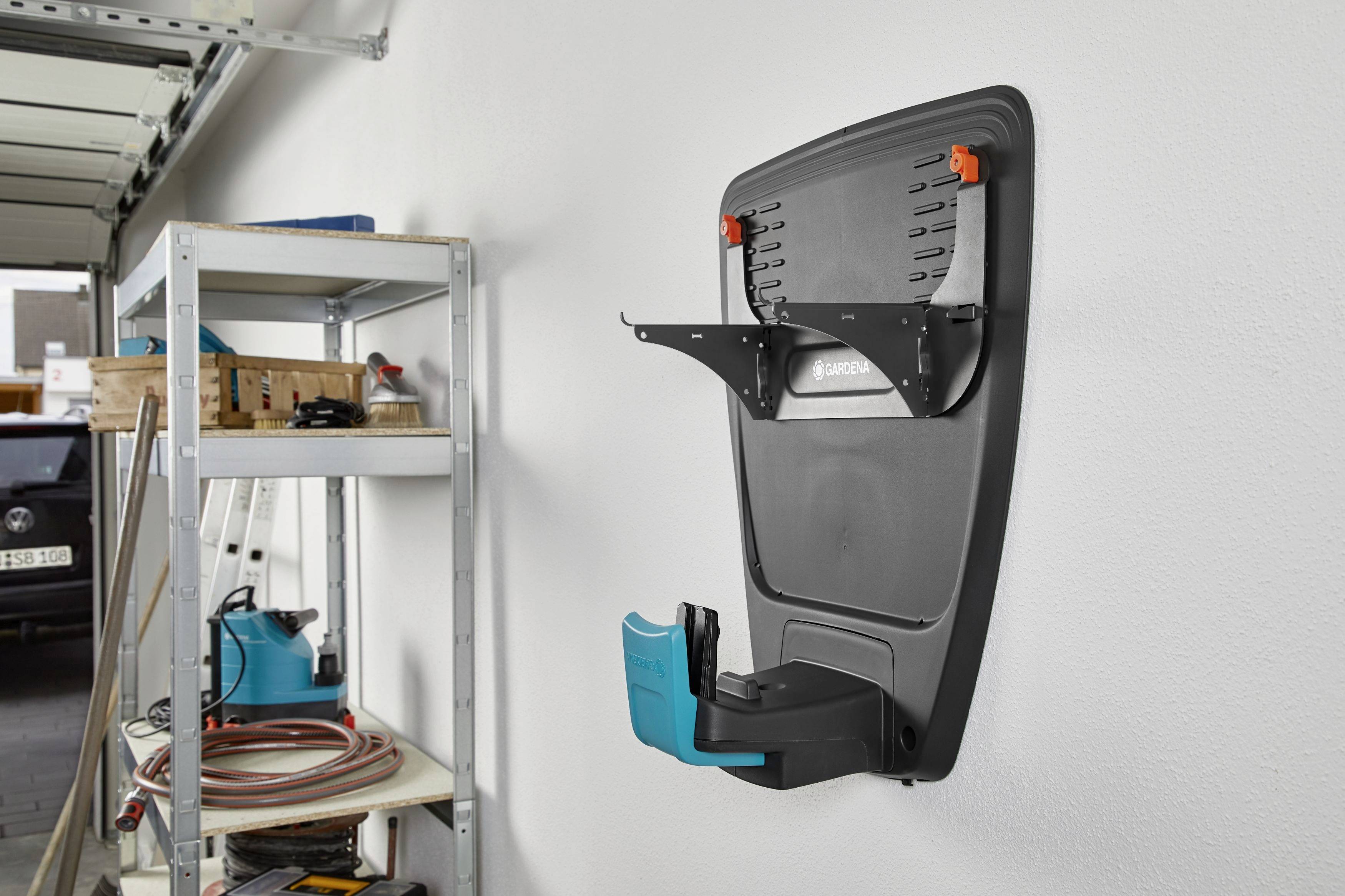 Na ścianie garażu widać zamontowaną ładowarkę z mocowaniem kabla. Po lewej stronie stoi regał z narzędziami i rolkami kabli.