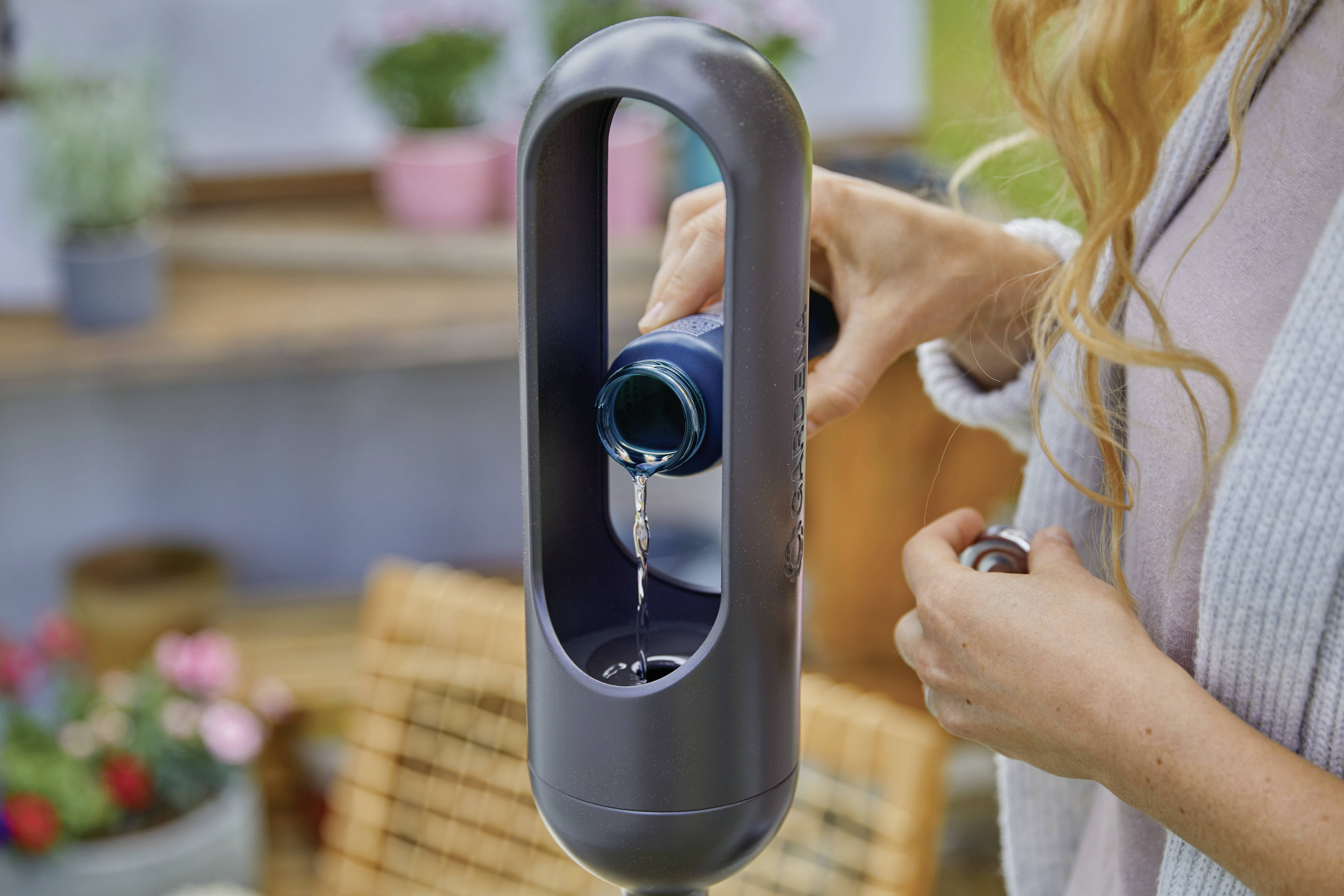 Osoba wlewa płyn z butelki do nowoczesnego środka czyszczącego ustawionego na zewnątrz. W tle widoczne są rośliny i nieostre meble ogrodowe.