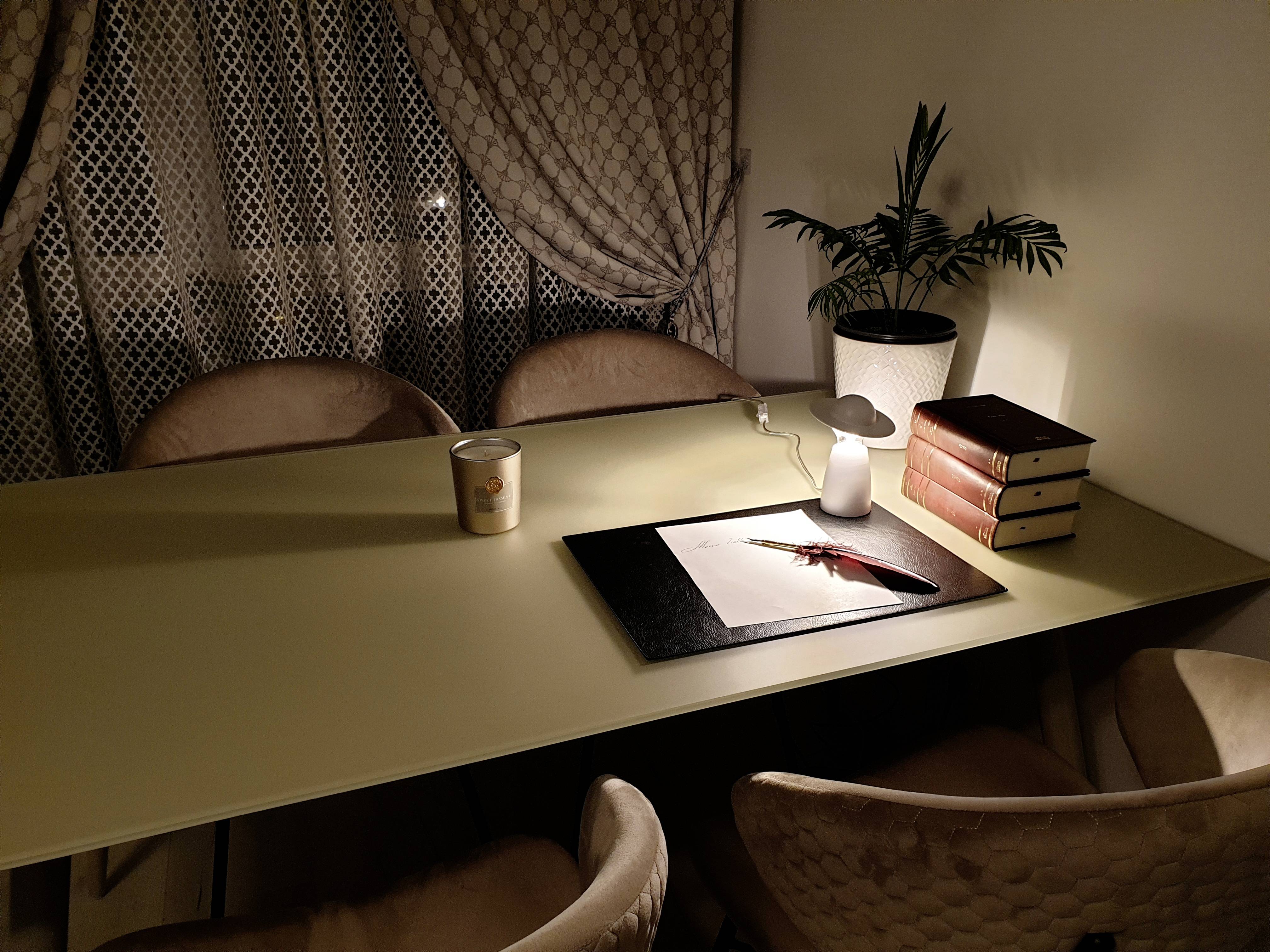 Stół z krzesłem, oświetlony lampą. Na stole leżą książki, filiżanka i obraz z piórem wiecznym.