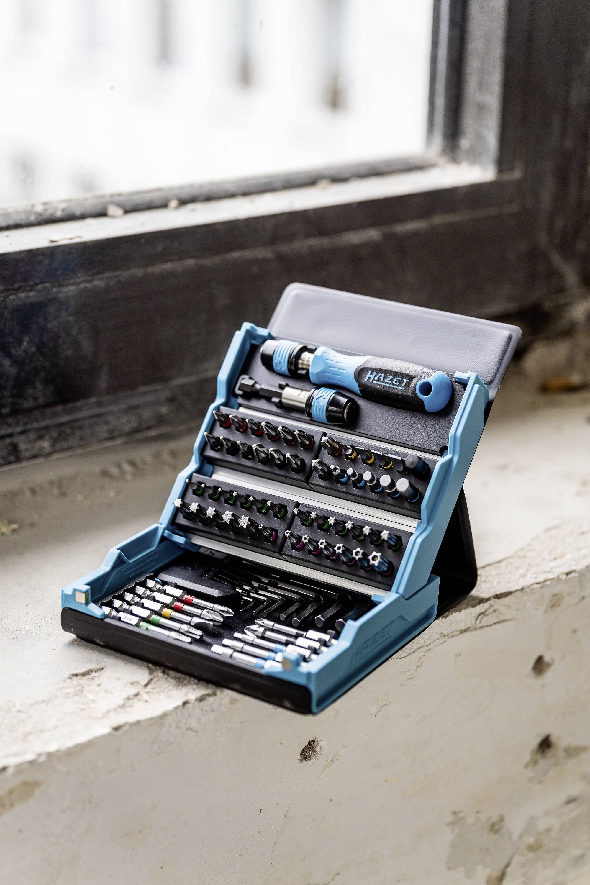 Zestaw różnych grotów śrubokrętowych z uchwytem leży w otwartej skrzynce narzędziowej na parapecie.