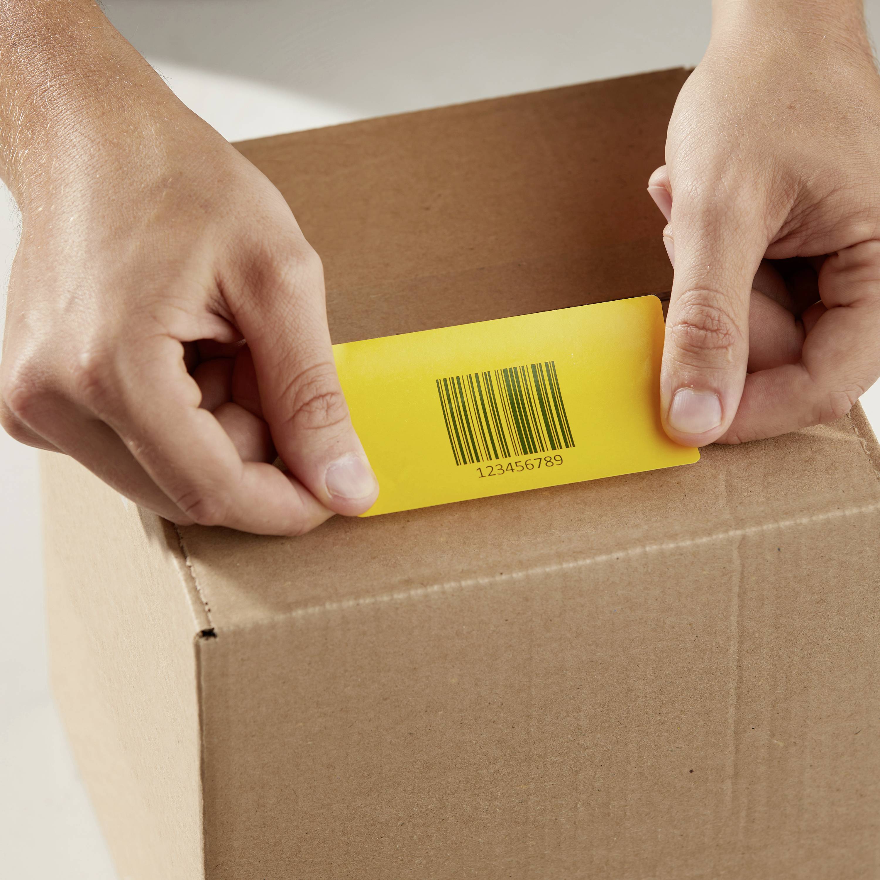 Ręce trzymają żółtą kartę z kodem kreskowym '123456789', którą wkłada się do brązowego kartonu.