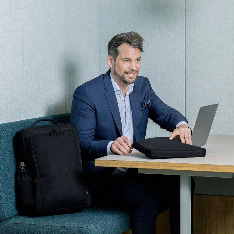 Mężczyzna w niebieskim garniturze siedzi w nowoczesnej przestrzeni biurowej przy stole, obok niego leży czarny plecak. Wygląda sympatycznie.