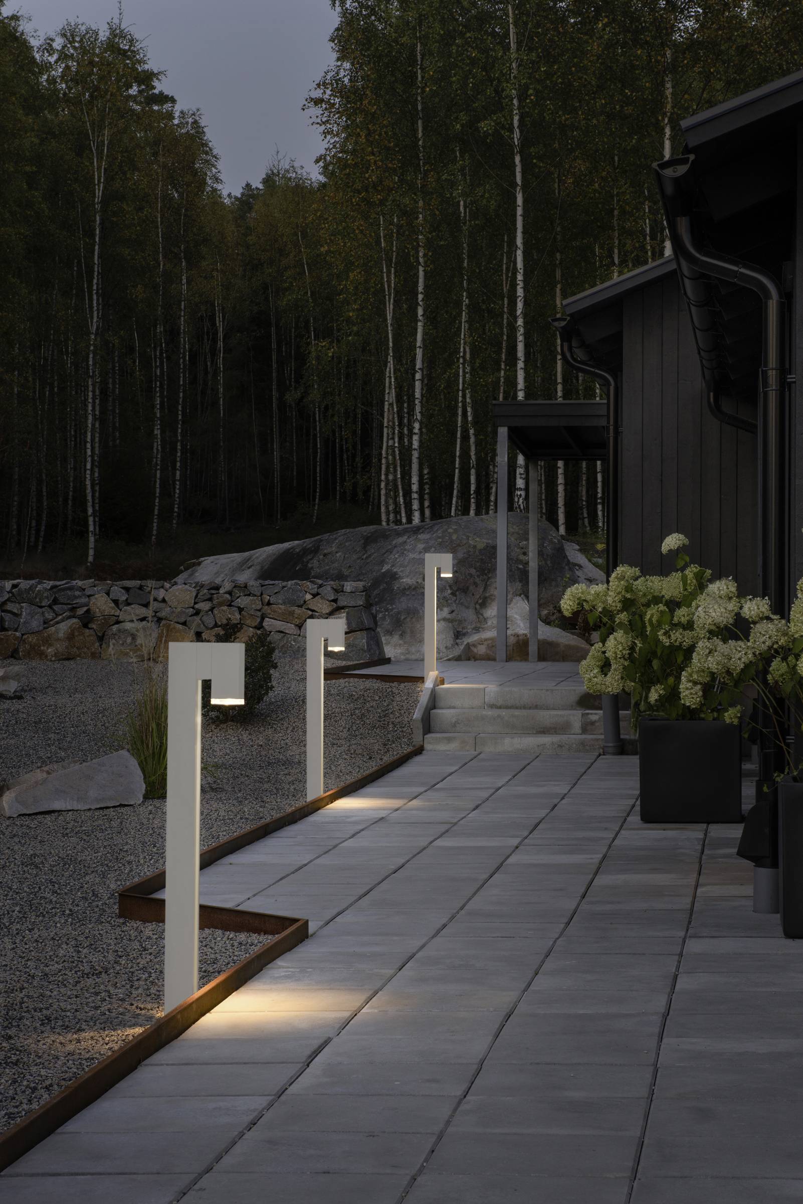 Oświetlona alejka ogrodowa o zmierzchu, z nowoczesnymi latarniami wyznaczającymi trasę wzdłuż nowoczesnego domu, przy którym stoją donice z kwiatami.