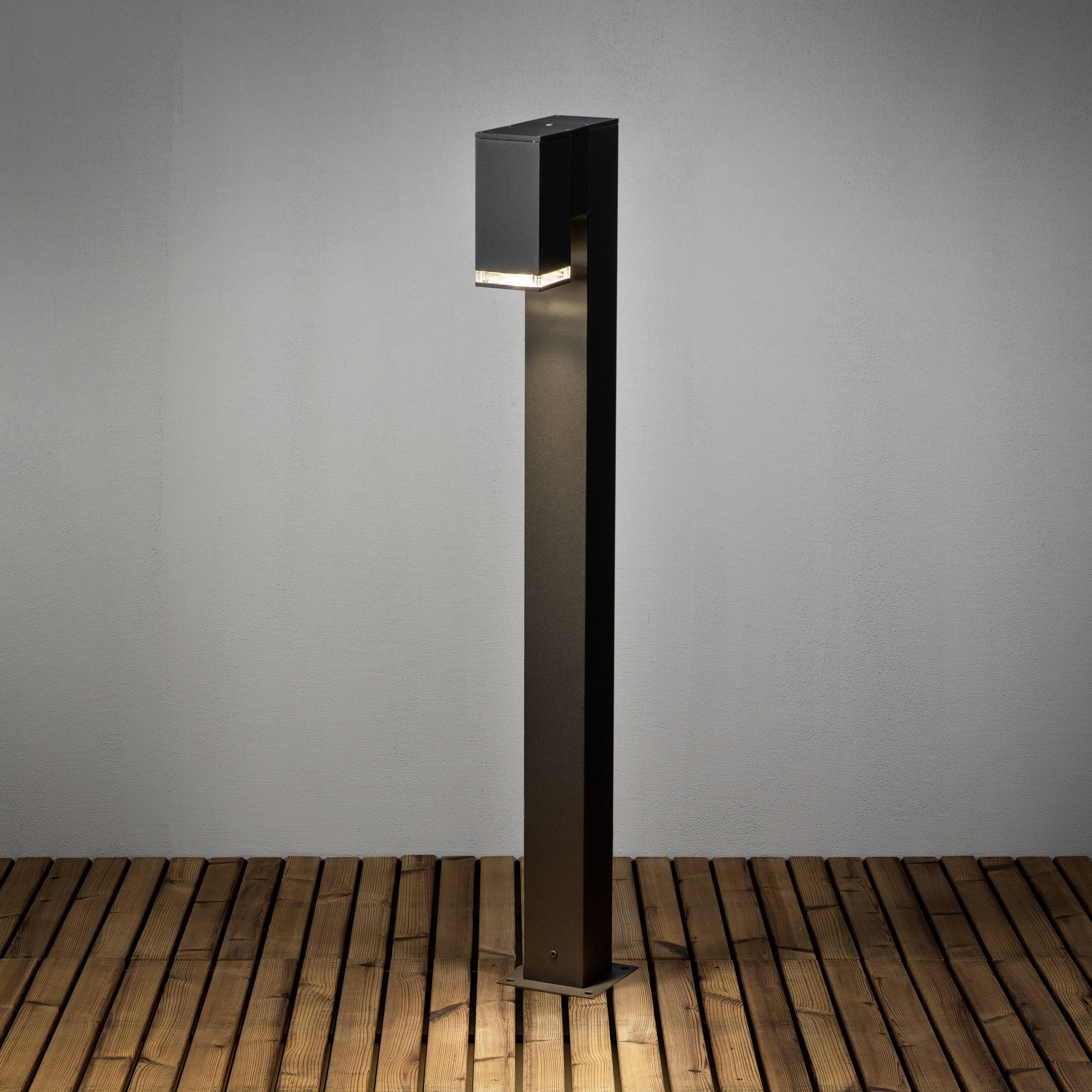 Nowoczesna, czarna lampa zewnętrzna rozświetla ciepłym światłem drewnianą podłogę.