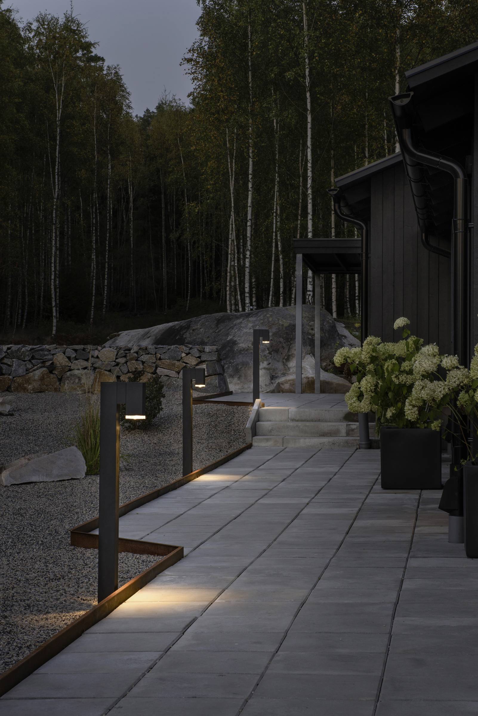 Wybrukowany taras z nowoczesnymi lampami zewnętrznymi oświetlającymi ścieżkę. W tle widoczne są drzewa i minimalistyczny dom.