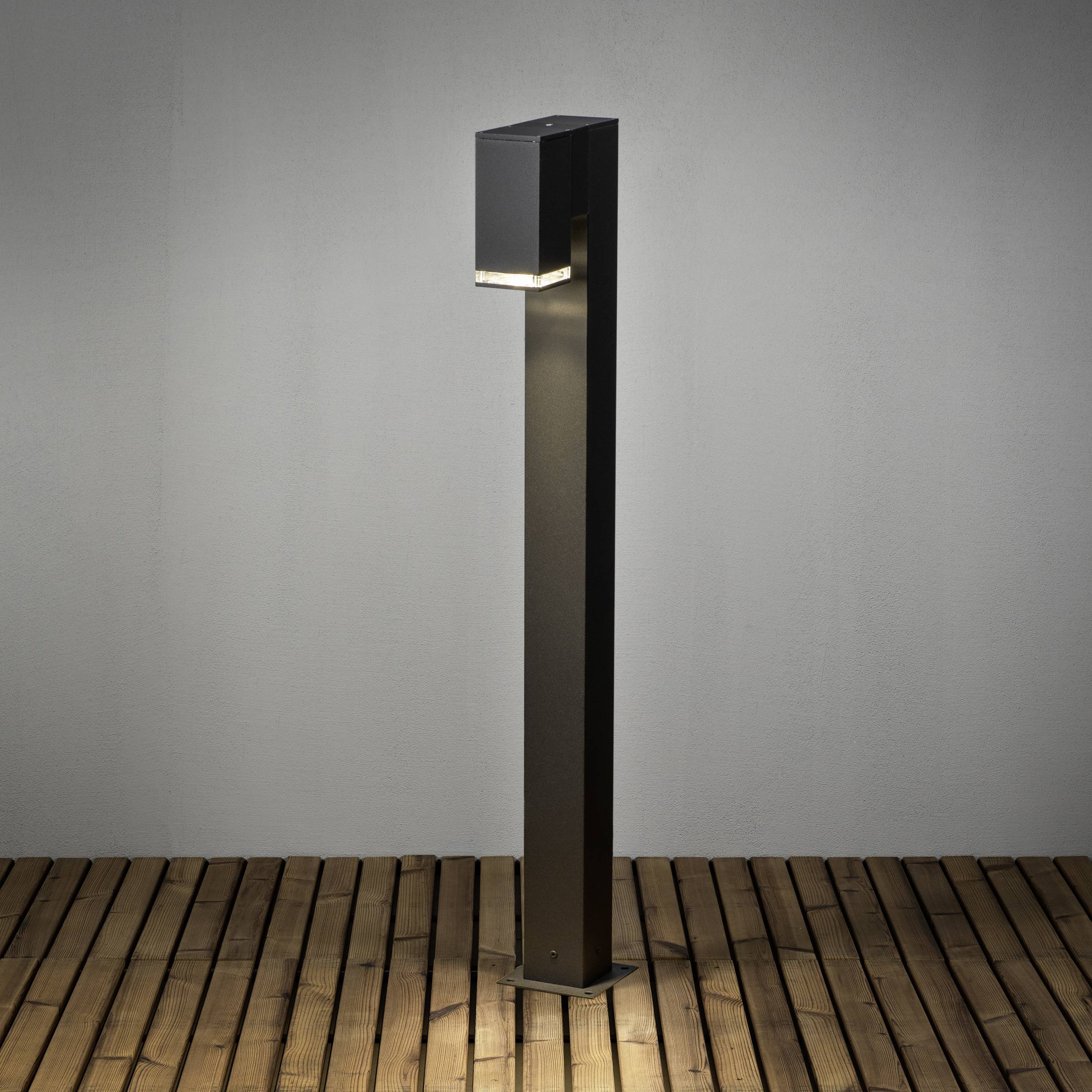 Czarna lampa zewnętrzna o nowoczesnym designie stoi na drewnianym tarasie i delikatnie oświetla otoczenie.