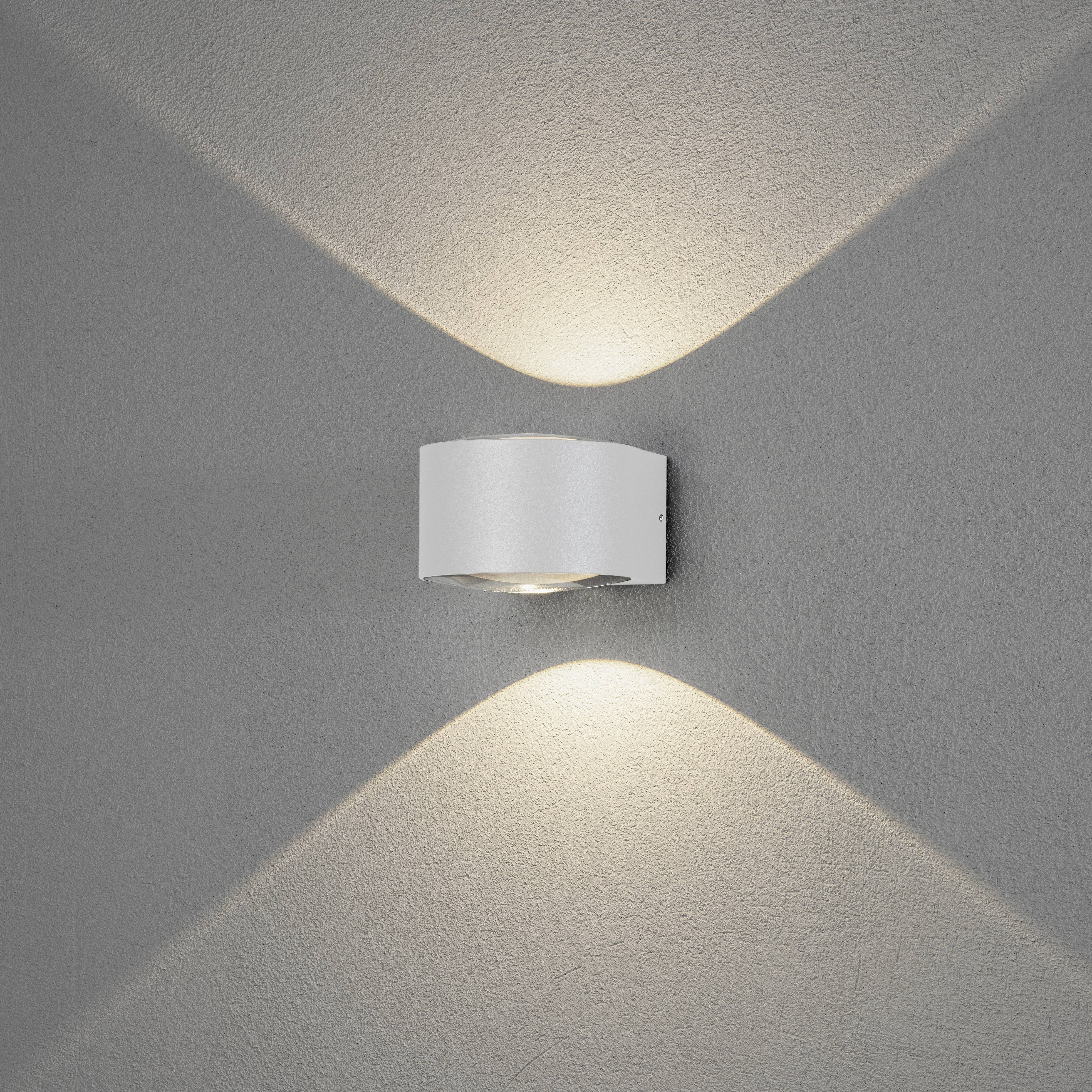 Biała lampa ścienna rzuca ciepłe światło w górę i w dół na szarą ścianę, tworząc elegancki, symetryczny wzór.