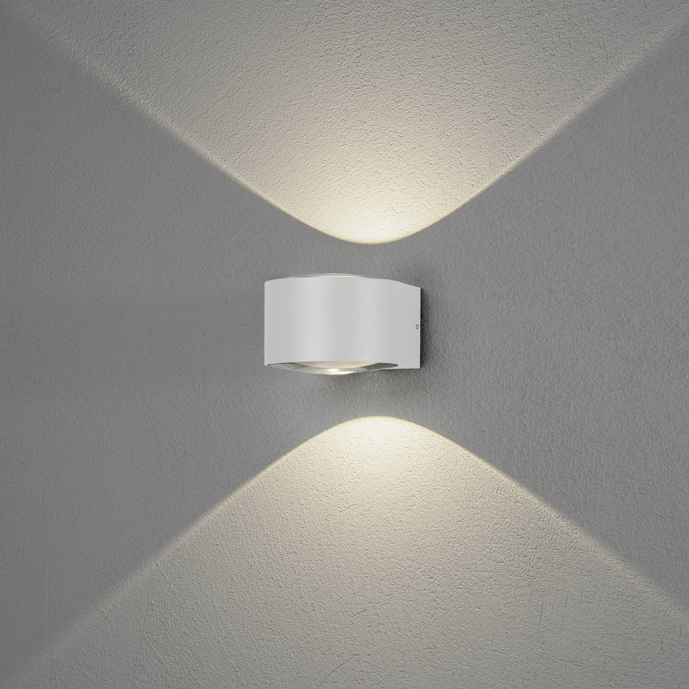 Nowoczesna lampa ścienna z metalu w minimalistycznym pomieszczeniu. Oprawa rozświetla przestrzeń miękkim światłem skierowanym w górę i w dół, tworząc elegancki klimat.