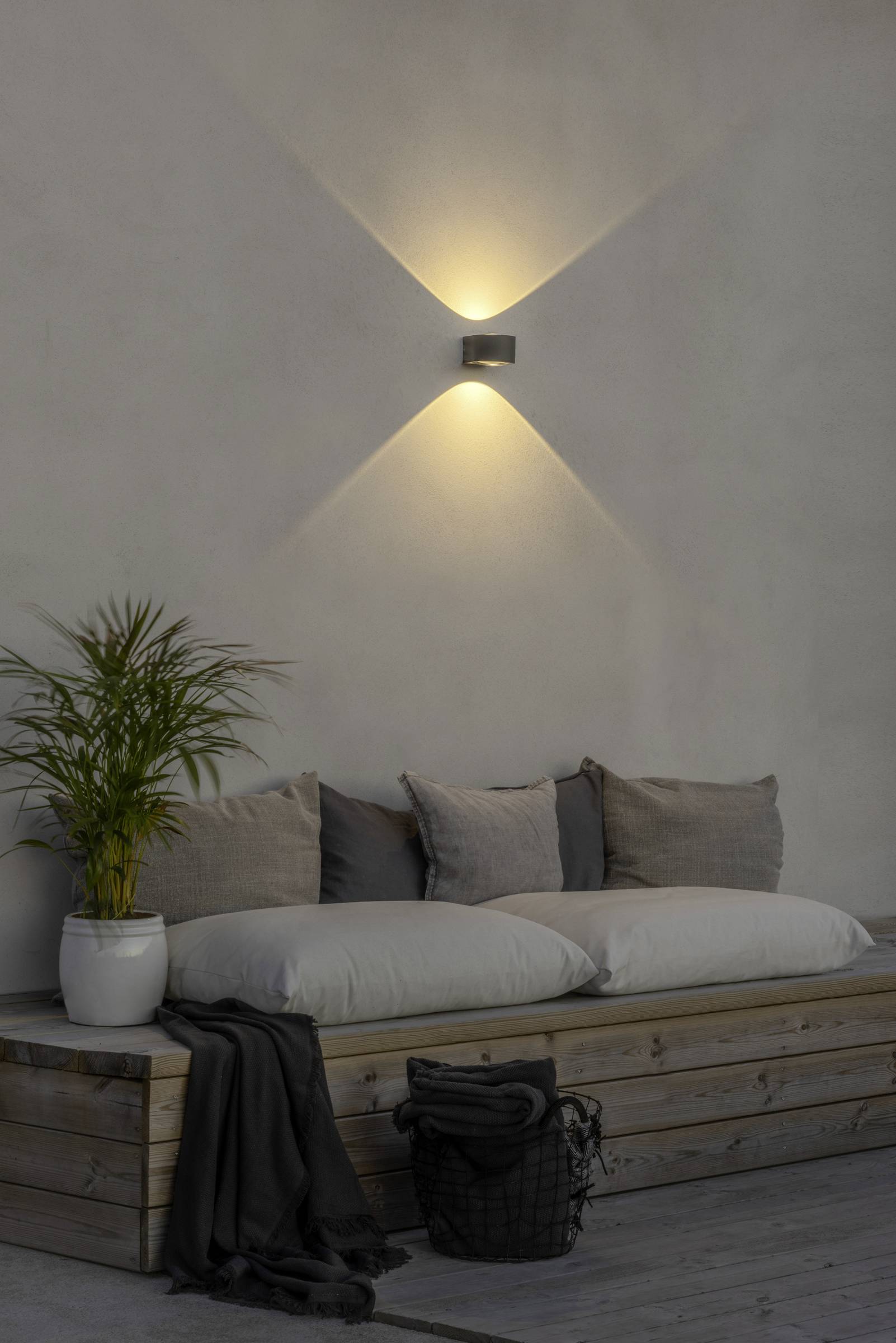 Minimalistyczny salon z drewnianą sofą, beżowymi poduszkami, doniczką z rośliną oraz nowoczesną lampą ścienną emitującą ciepłe światło.