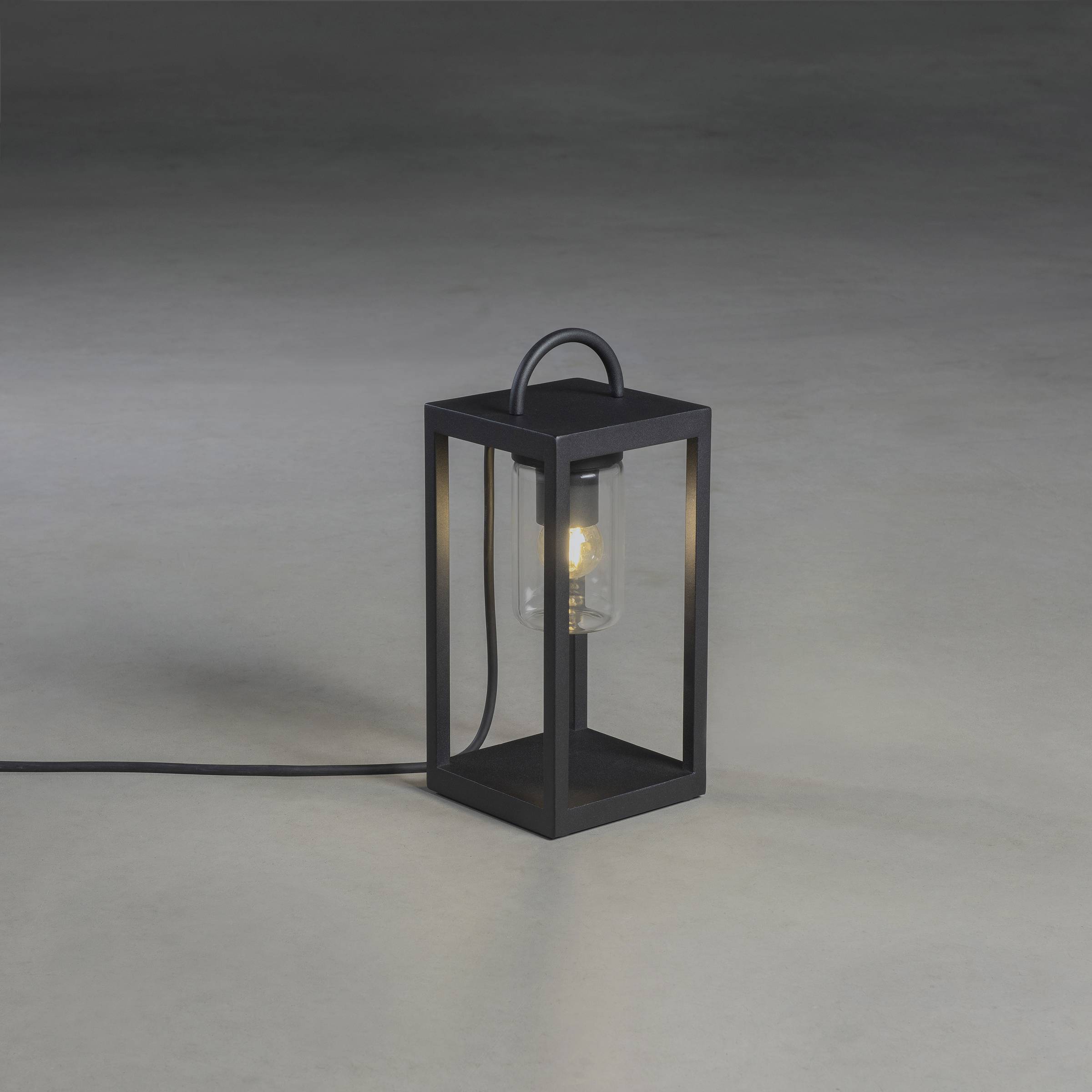 Czarna lampa stołowa z żarówką, osadzona w minimalistycznej metalowej ramie, stojąca na szarym podłożu. Kabel widoczny na podłodze.
