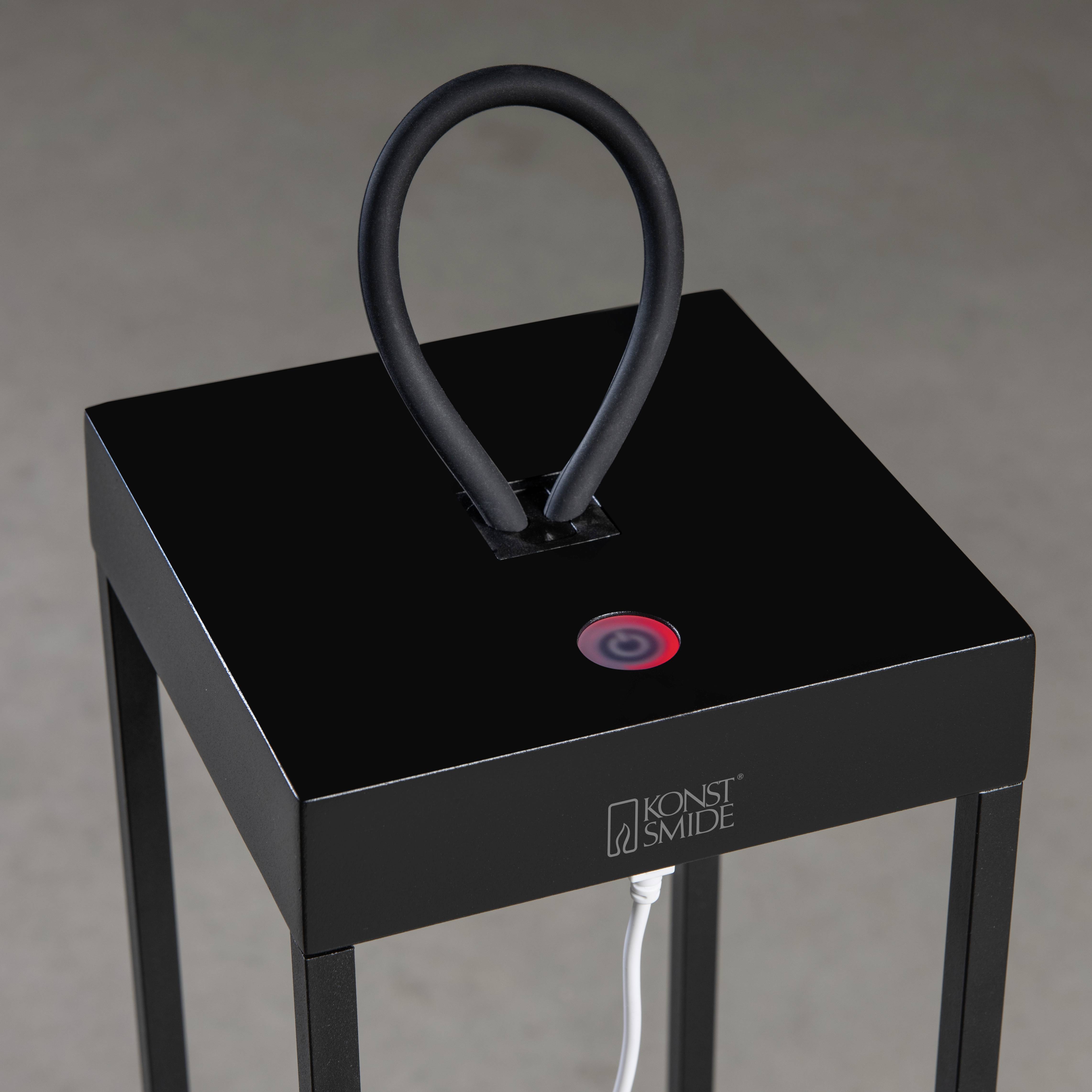 Czarna, minimalistyczna lampa z kwadratową podstawą i wygiętym łukiem, na którym widnieje logo 'Konst Smide'.
