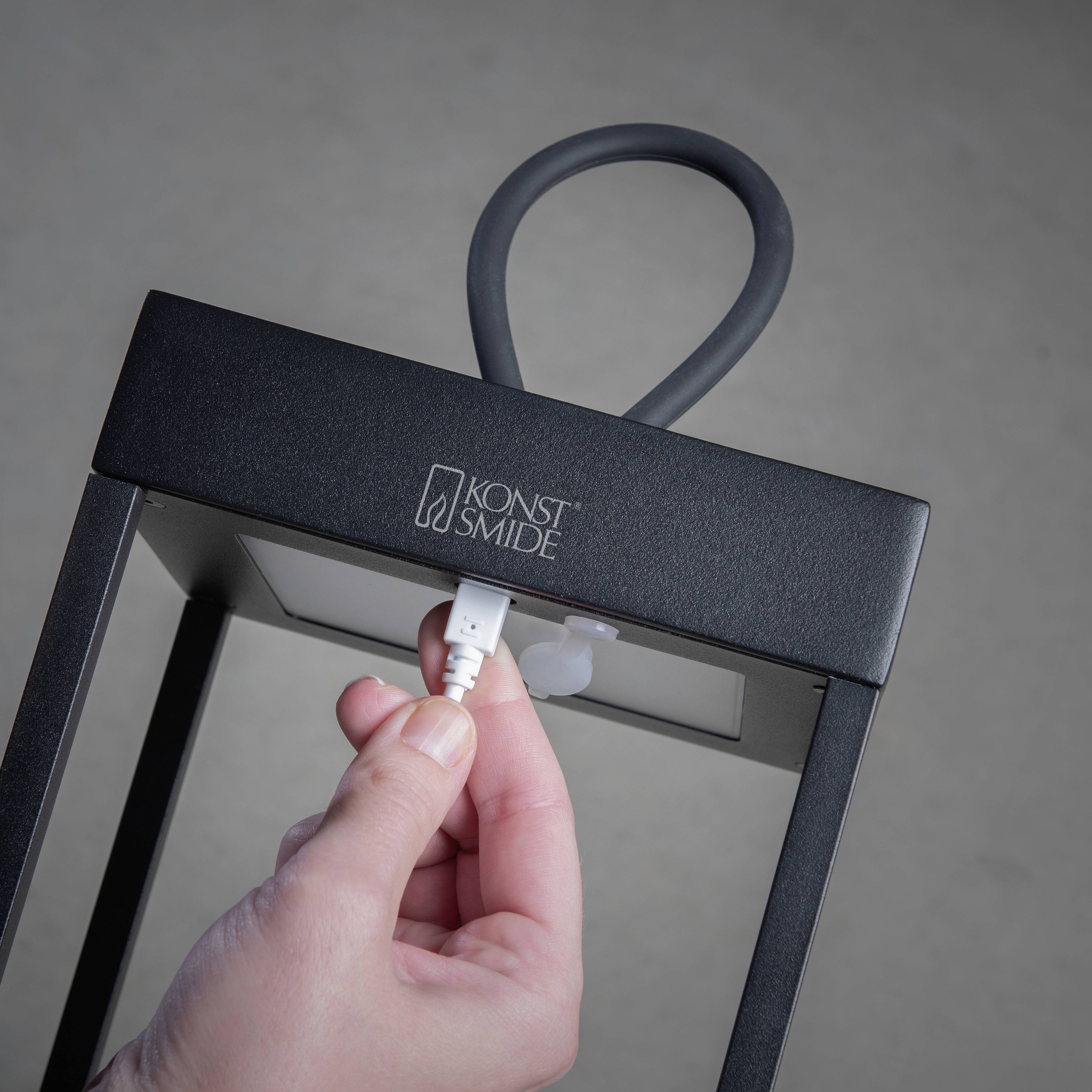 Ręka wkłada przewód ładujący do czarnej stołowej lampy. Na górze widoczne jest logo 'Konst Smide'.