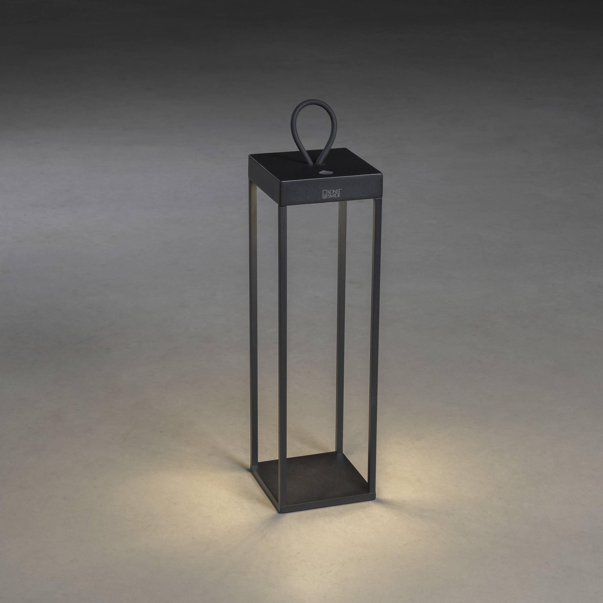Nowoczesna, prostokątna latarnia z czarnego metalu, stojąca na ciemnym podłożu i delikatnie rozświetlająca przestrzeń od dołu.