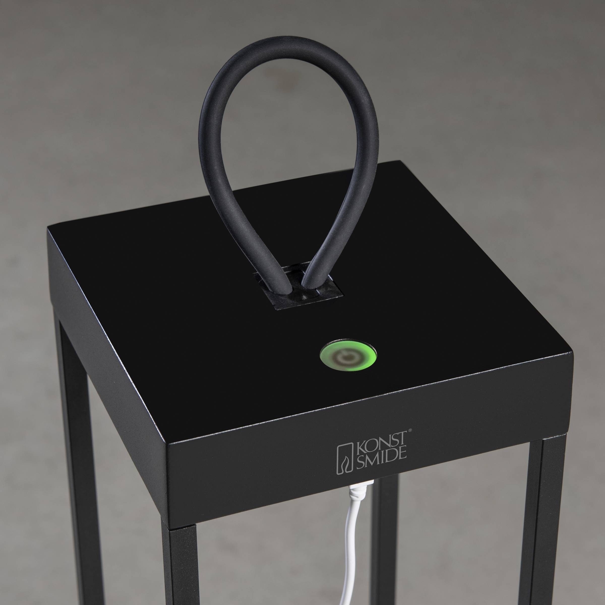 Kwadratowy, czarny, nowoczesny obiekt designerski do użytku zewnętrznego z okrągłą lampą LED na górze i uchwytem w kształcie pierścienia; stoi na czterech nogach.