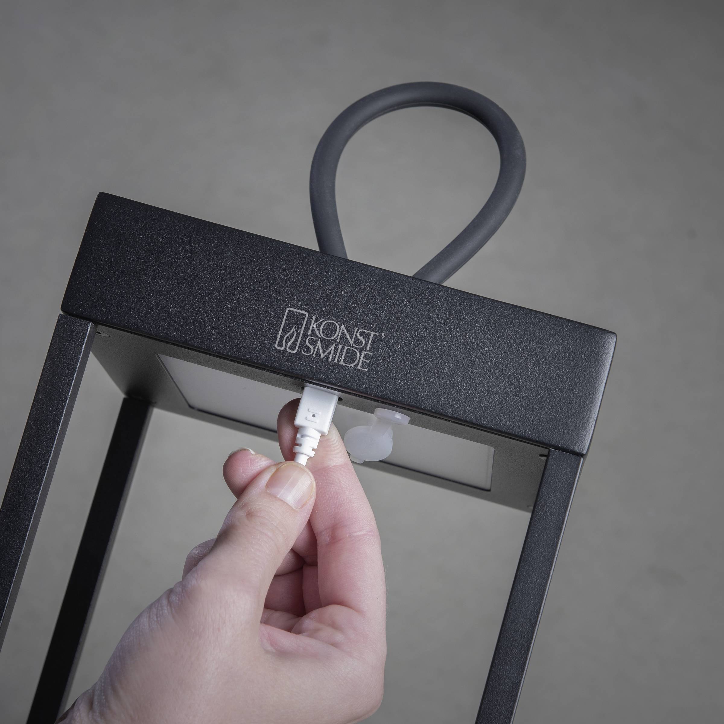 Osoba wkłada białą wtyczkę kabla do czarnej latarni o kwadratowej ramie, otwartej od góry.