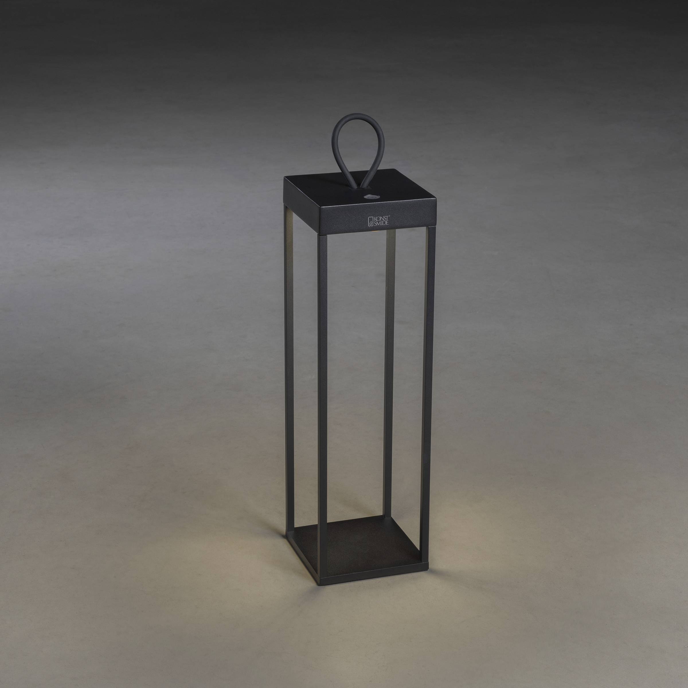 Minimalistyczny, czarny lampion w nowoczesnym stylu stoi na szarym podłożu i słabo świeci w środku.