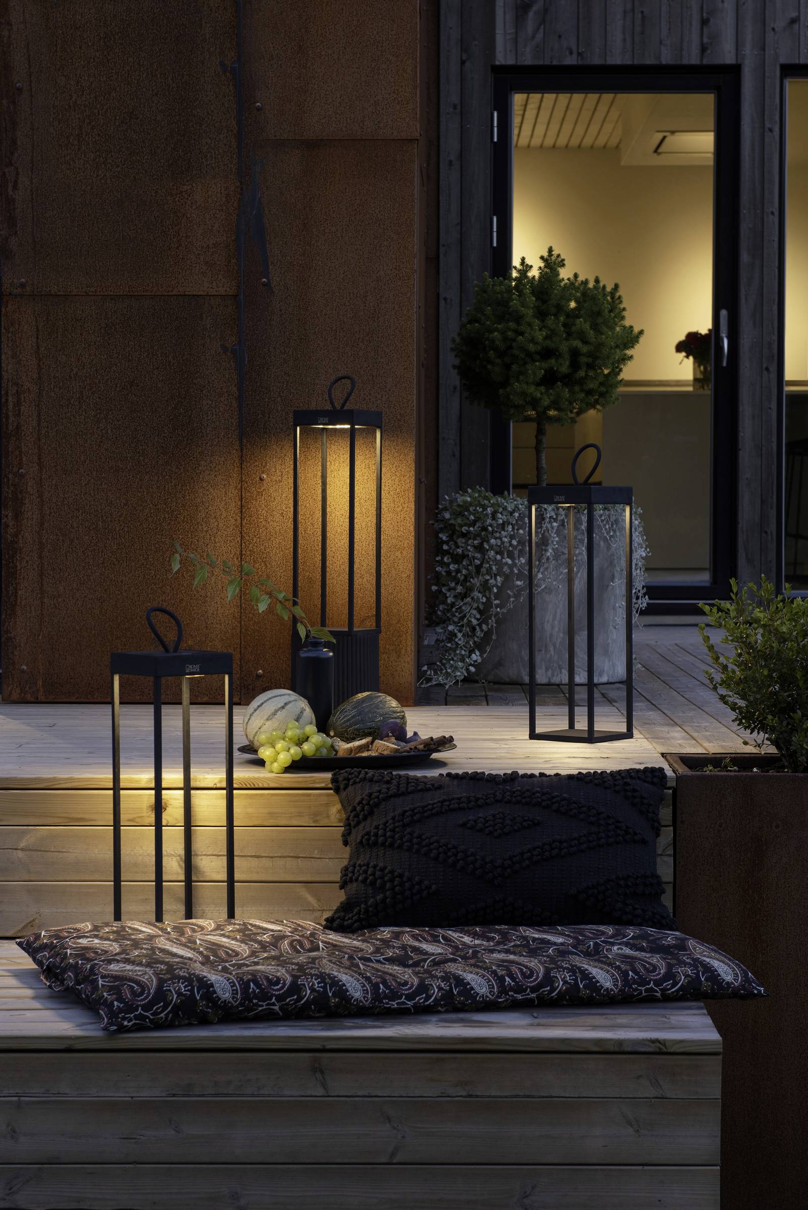 Przestrzeń zewnętrzna z kilkoma oświetlonymi latarniami na drewnianych stopniach, otoczona roślinami i dyniami, przed oświetlonym domem.