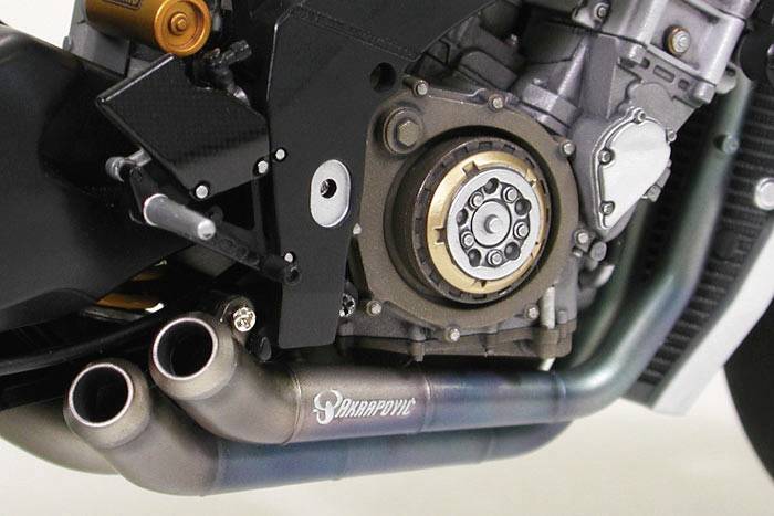 Zbliżenie silnika motocyklowego, przedstawiające układ wydechowy i komponenty silnika. Na jednym z końcowych elementów układu wydechowego widoczna jest marka 'Akrapovic'.