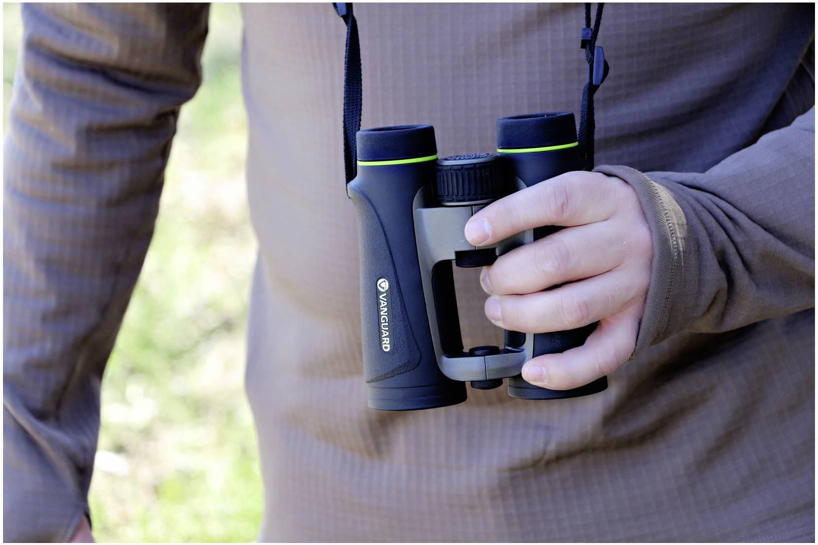 Osoba trzyma lornetkę w prawej ręce, która wisi na pasku przewieszonym przez szyję. Tło jest rozmyte.