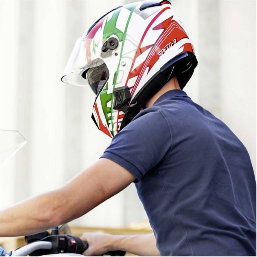 Osoba ubrana w niebieską koszulkę prowadzi motocykl, na głowie mając kolorowy kask w odcieniach czerwieni i zieleni.