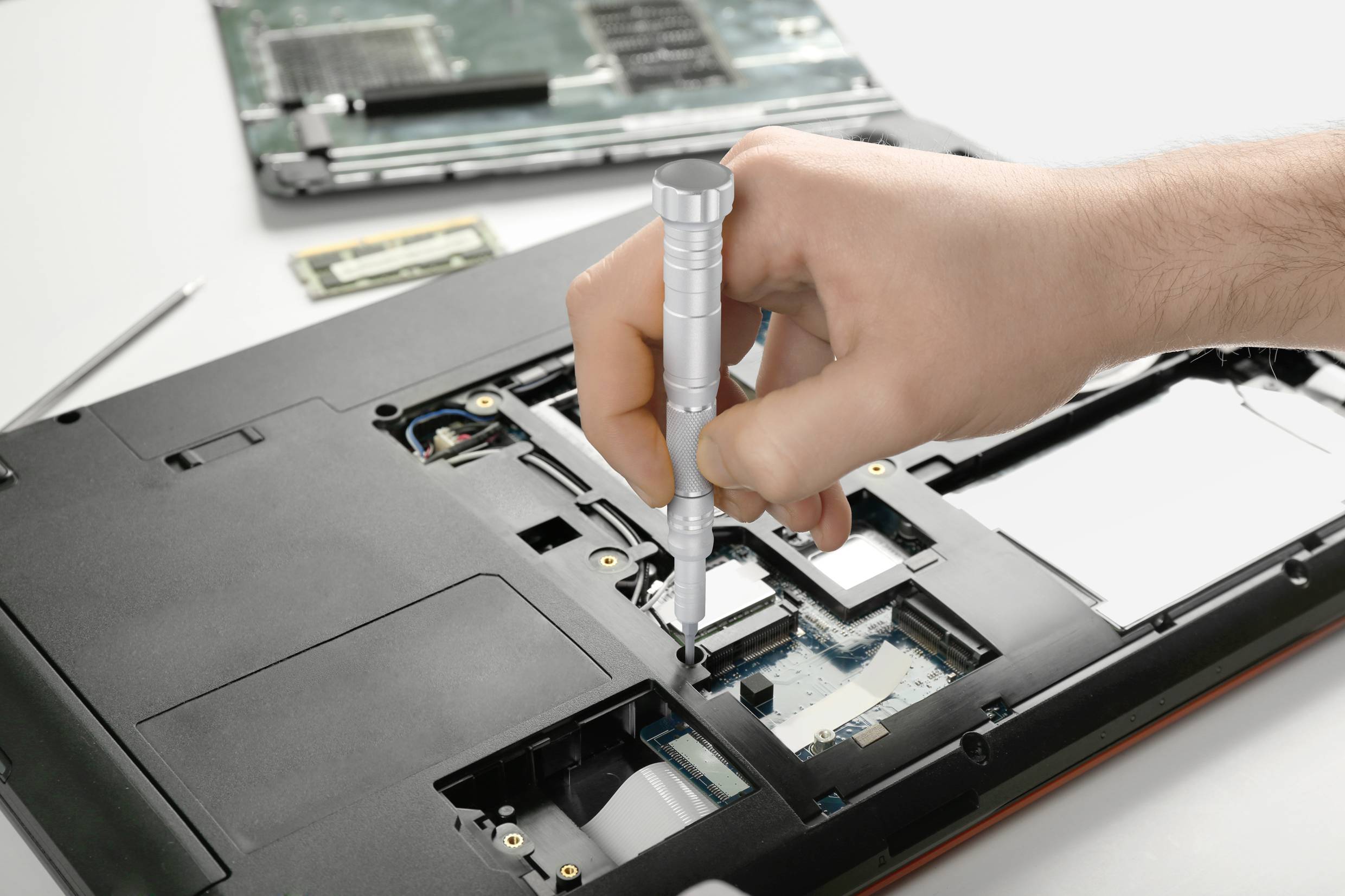 Ręka naprawia otwarte obudowy laptopa za pomocą śrubokręta. Widoczny jest moduł RAM oraz płyta główna.