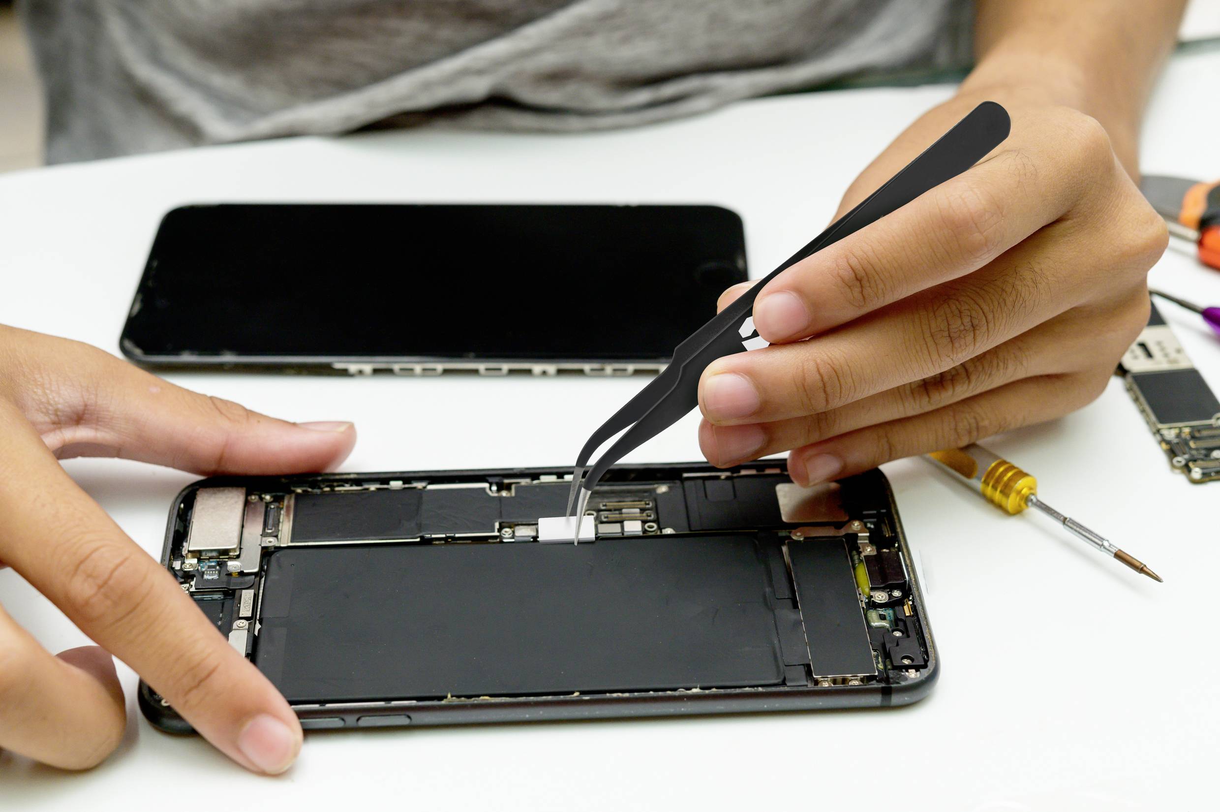 Osoba naprawia wnętrze smartfona za pomocą pęsety. Na stole leży kolejna rama telefonu i narzędzia.