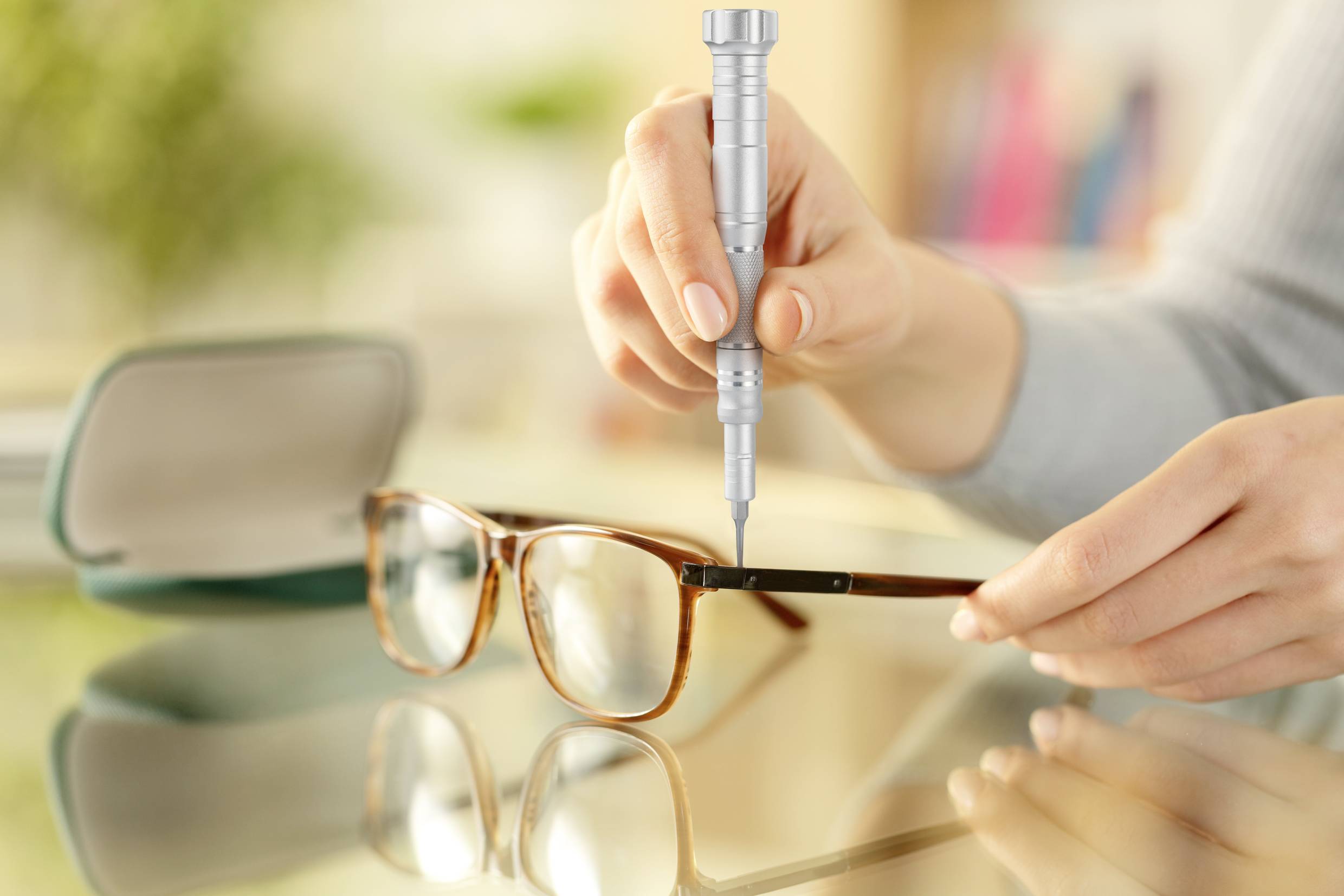 Osoba trzyma okulary i dokręca wkrętak śrubkę zawiasu. W tle widoczne jest etui na okulary.