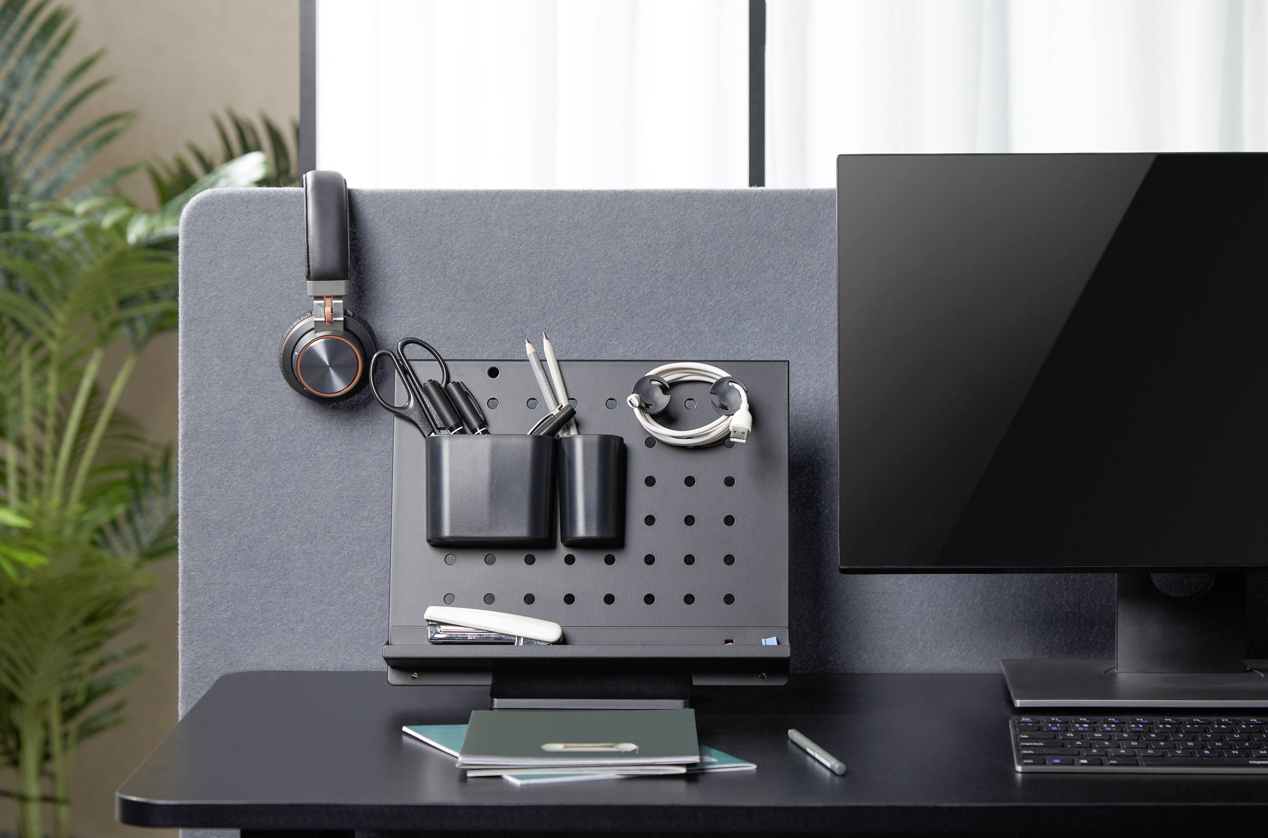 Stanowisko pracy z biurkiem, laptopem, słuchawkami i organizatorem na kable i długopisy. W tle rośliny i jasne okno.