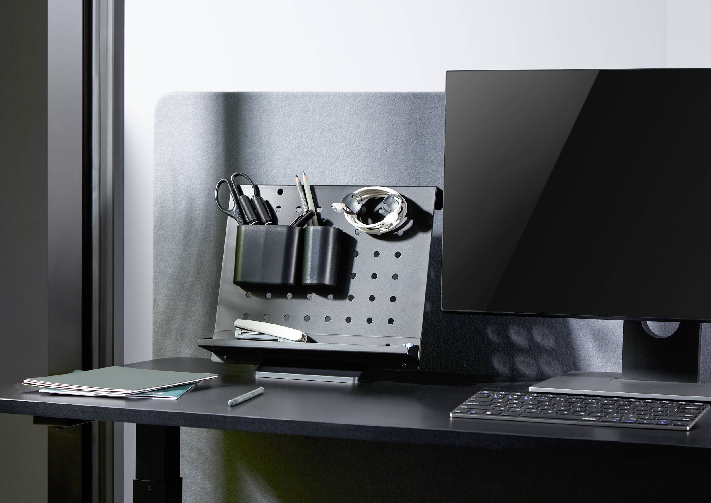 Uporządkowane biurko z monitorem, klawiaturą, notatnikiem, długopisem i tablicą organizacyjną z przyborami do pisania i słuchawkami w uchwytach.