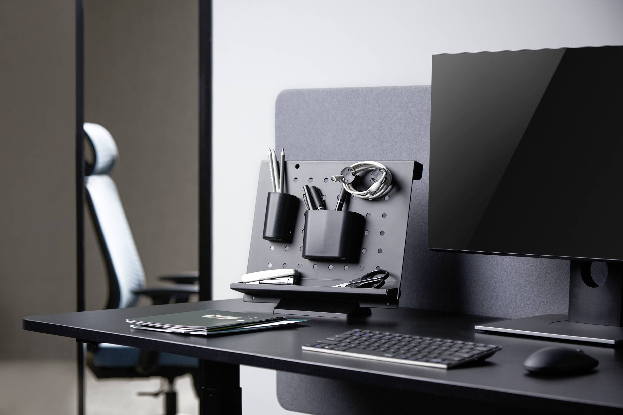 Nowoczesne stanowisko pracy z monitorem, klawiaturą, notatnikiem i pojemnikami na długopisy na czarnym stole. W tle stoi krzesło biurowe.