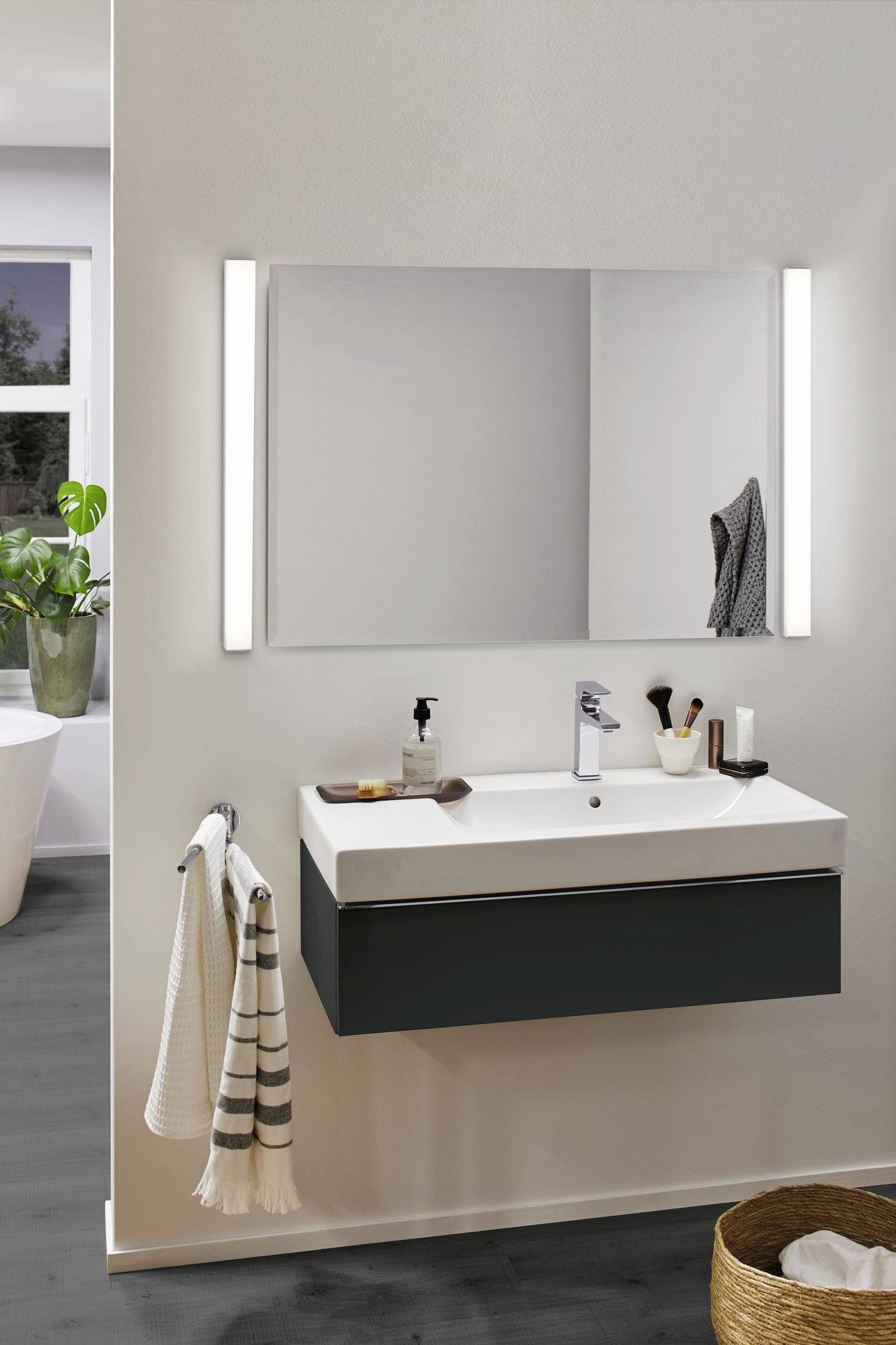 Nowoczesna łazienka z umywalką, dużym lustrem i ręcznikami; po lewej stronie okno z roślinami; po prawej stronie kosz.