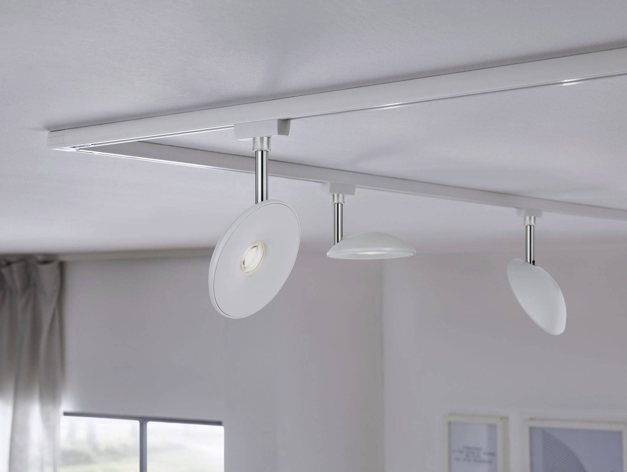 Trzy nowoczesne, białe reflektory sufitowe na szynie, jeden z nich świeci. W tle okna i ramki do zdjęć. Minimalistyczny design.