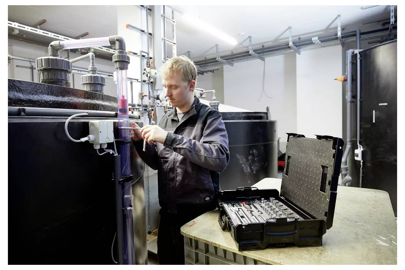 Osoba w warsztacie reguluje urządzenie na rurze obok dużych zbiorników. Otwarta walizka z narzędziami znajduje się na pobliskiej ławce.