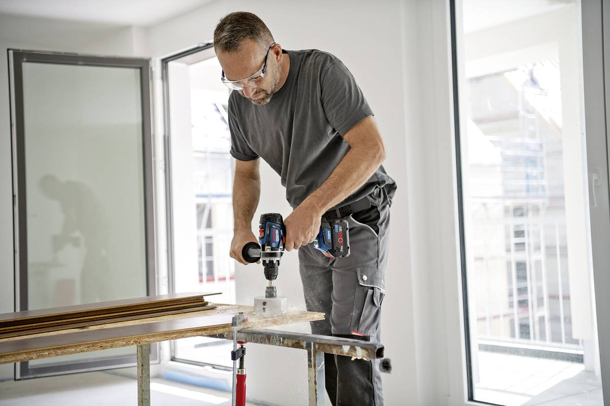 Mężczyzna pracuje z wiertarką przy stole w jasnym pomieszczeniu z dużymi oknami. Na sobie ma okulary ochronne i profesjonalne ubranie robocze.