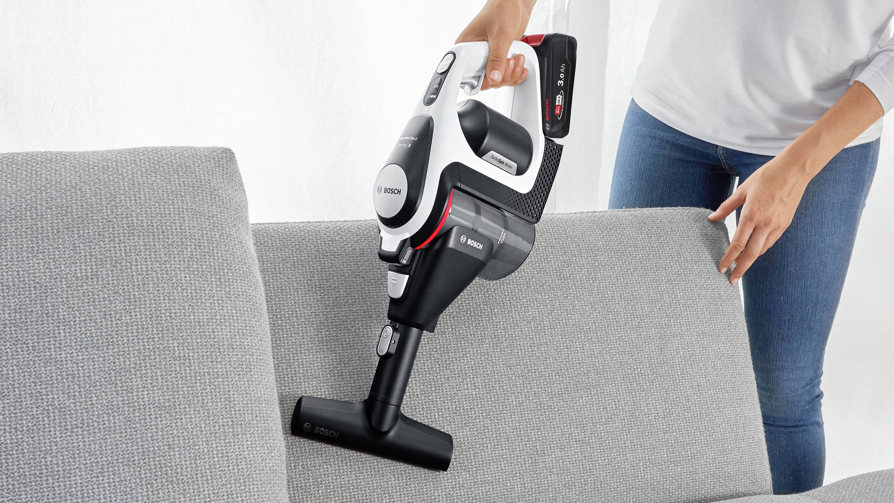 Osoba używa bezprzewodowego odkurzacza do czyszczenia szarej kanapy, prezentując wygodne sprzątanie mebli.