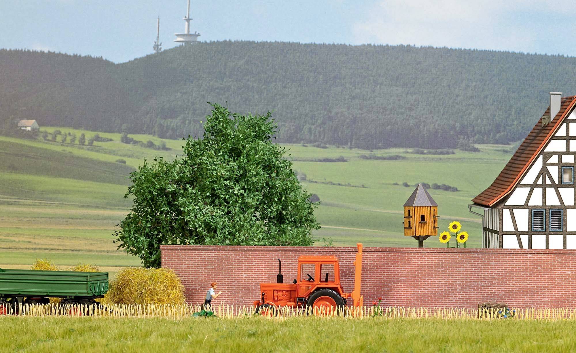 Miniaturowa diorama krajobrazowa: Traktor jadący wzdłuż muru, obok dom szachulcowy i karmnik, wzgórza w tle.