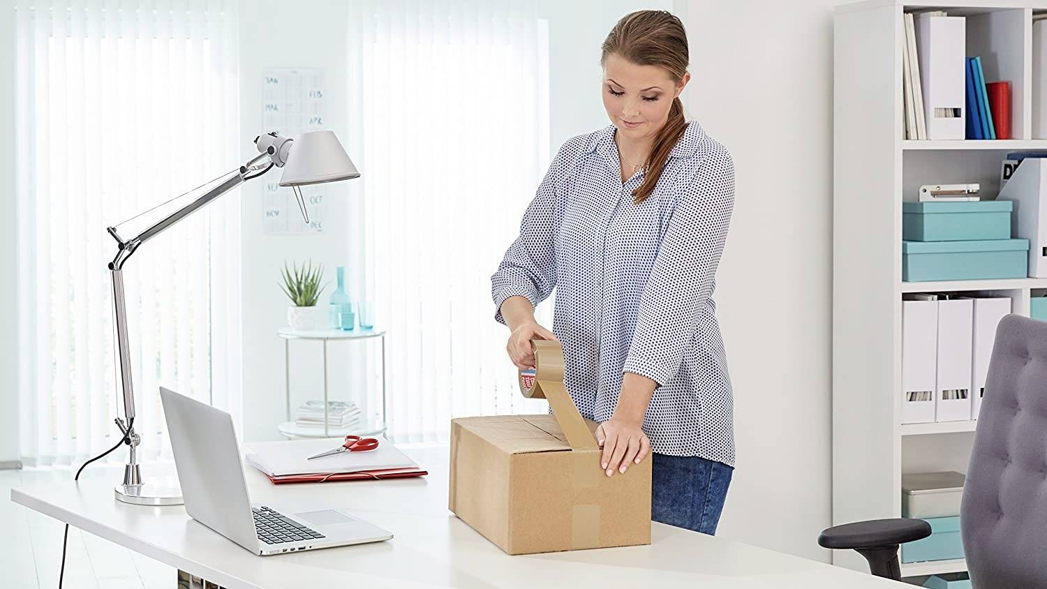 Kobieta w biurze uszczelnia karton taśmą klejącą. Na pierwszym planie widać biurko z lampą biurkową, laptopem i segregatorami w tle.