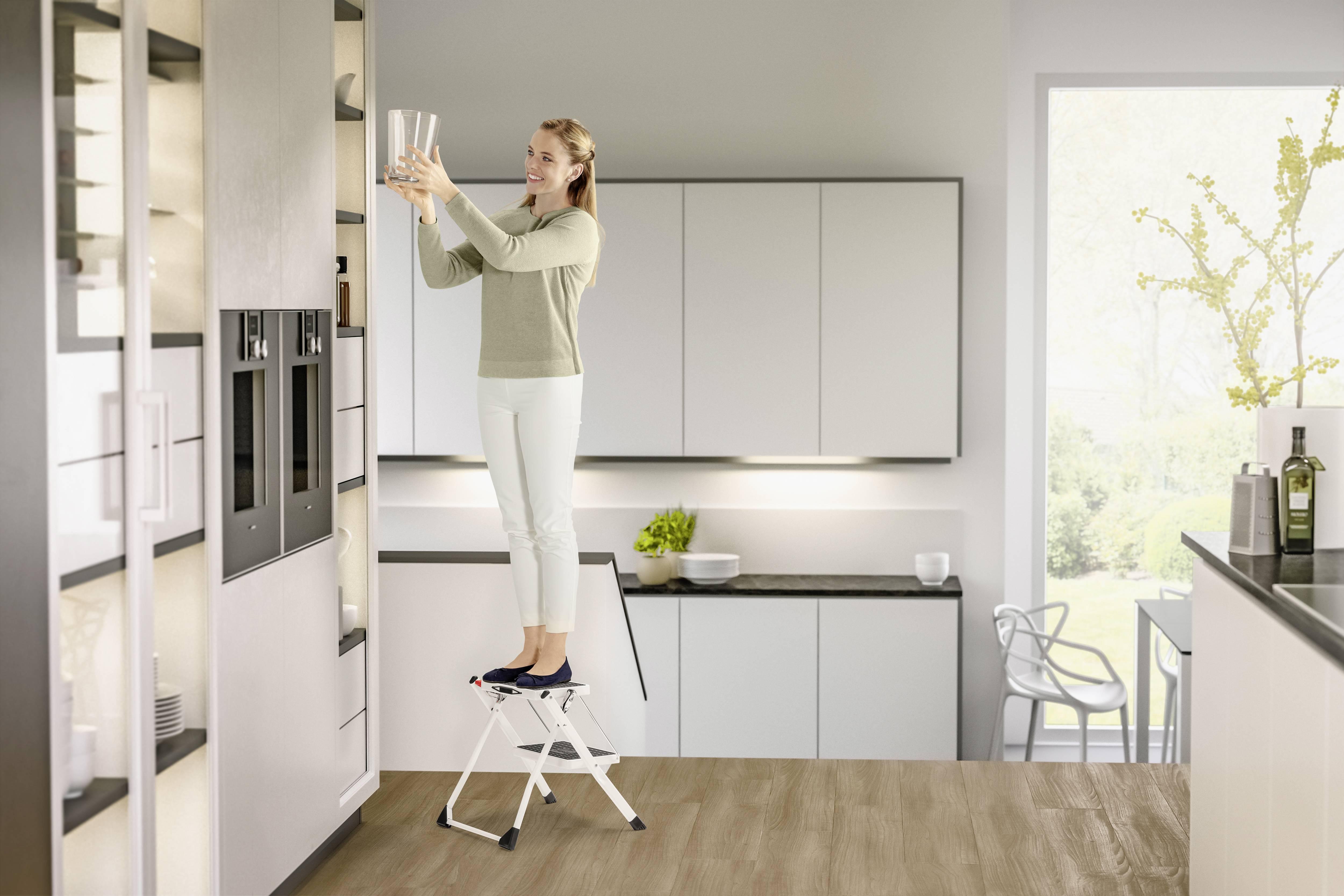 Kobieta stoi na taborecie i wkłada szklanki do górnej półki nowoczesnej kuchni. Ma na sobie jasne ubranie.