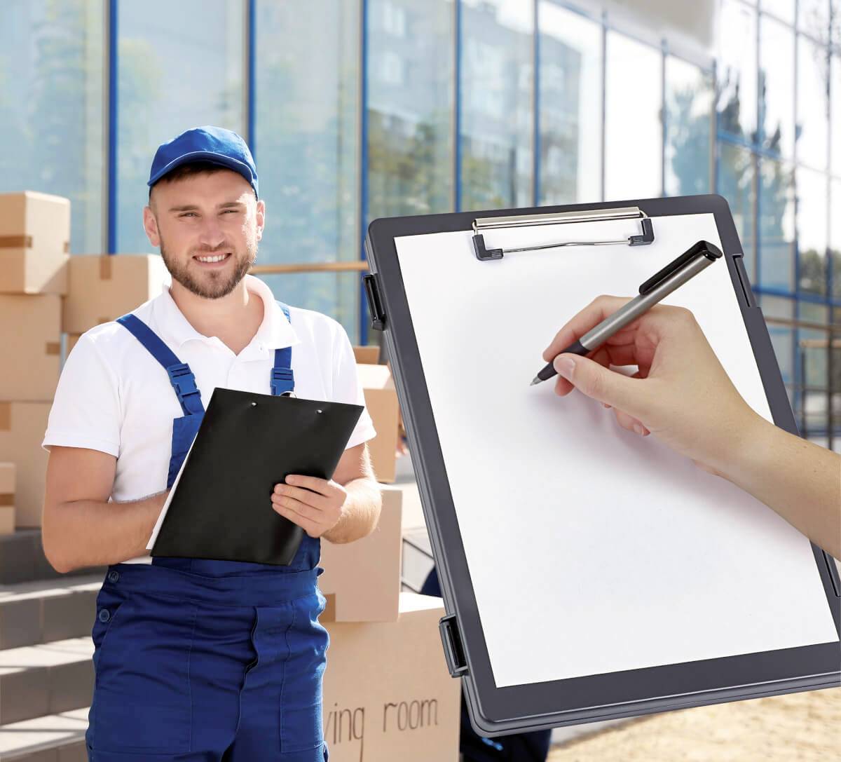 Mężczyzna w niebieskim kombinezonie roboczym i czapce stoi przed kartonami z dokumentami w ręku; jedna ręka jest zajęta pisaniem na kartce papieru.