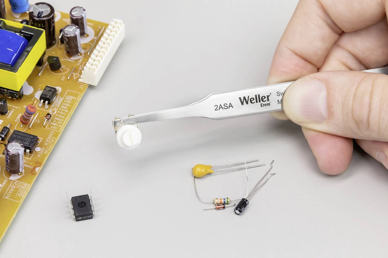 Ręka trzyma pęsetę z elementem elektronicznym nad płytką drukowaną. Obok leżą kolejne komponenty.