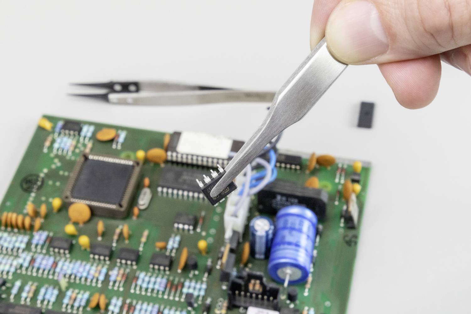 Dłoń trzyma za pomocą pincety mały mikrochip nad płytką drukowaną. W tle widoczne są komponenty elektroniczne.