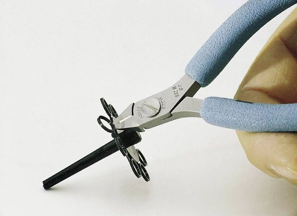 Dłoń trzyma niebieskie szczypce boczne, które przecinają czarny plastikowy przedmiot. Ostrze szczypców i precyzyjne cięcie są w centrum uwagi.