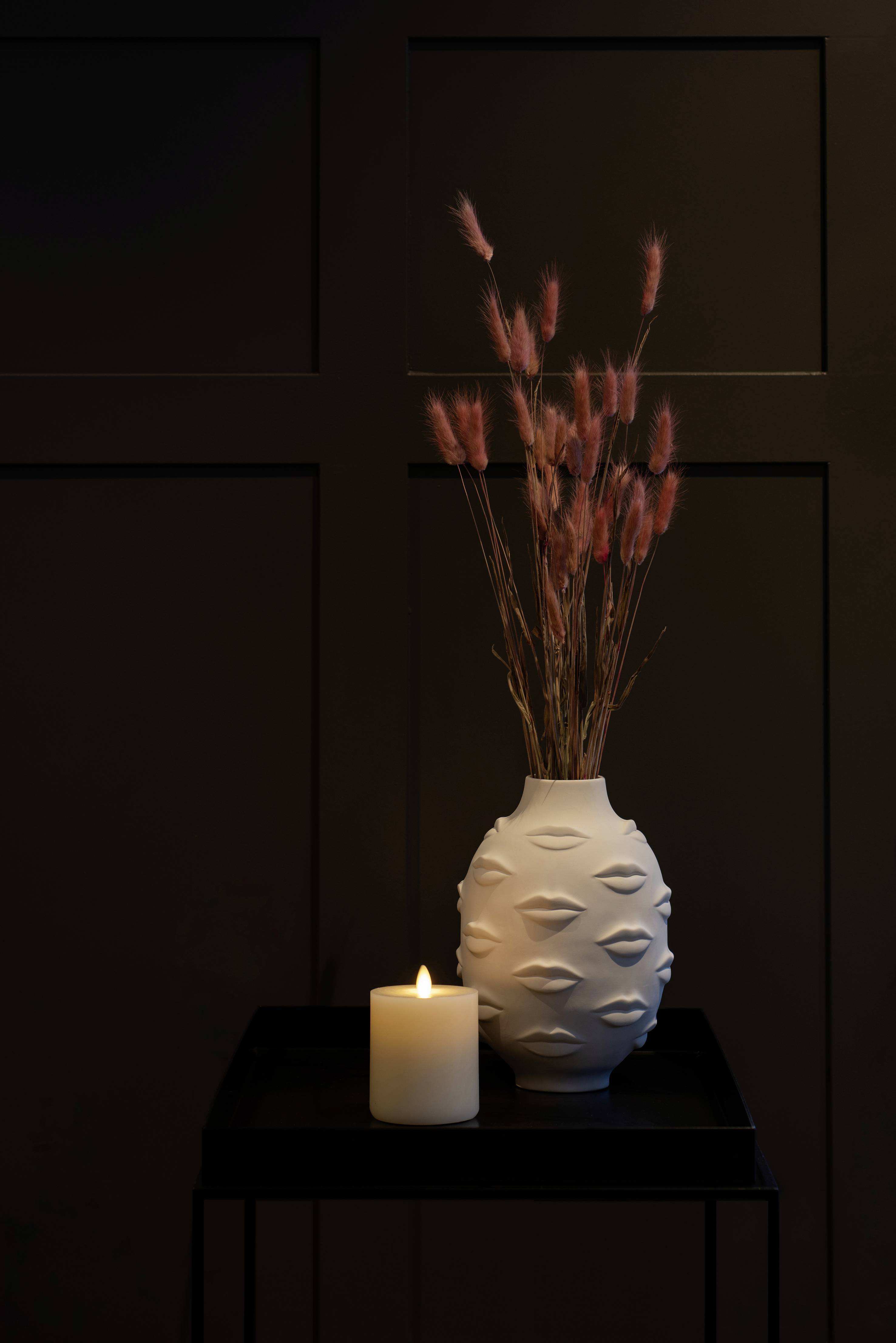 Biała waza z motywem ust i różowymi suszonymi kwiatami stoi na ciemnym stole obok płonącej świecy na ciemnym tle.