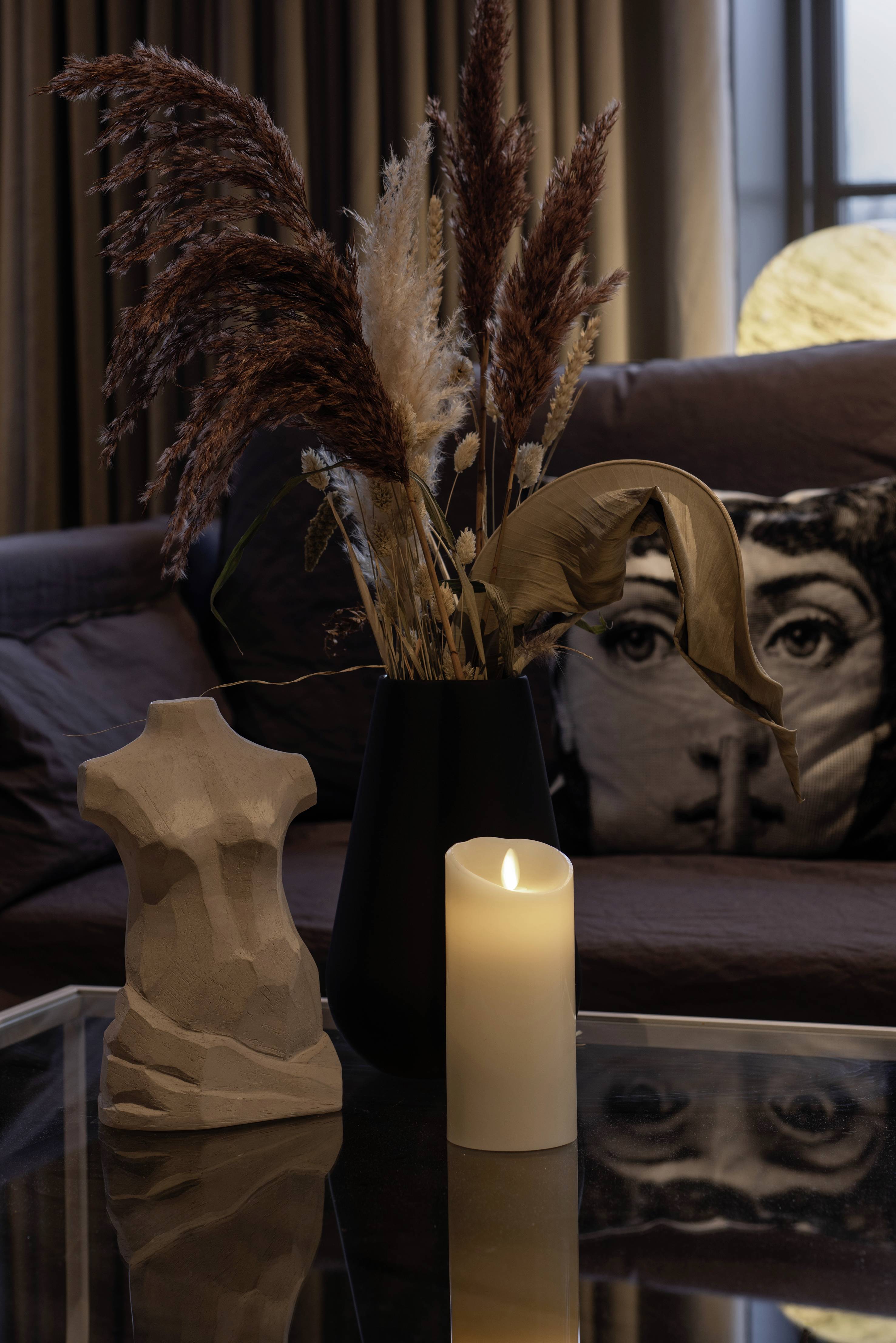 Światło świecy oświetla stół z abstrakcyjnym popiersiem, suchym bukietem kwiatów w wazonie oraz sofą w tle.
