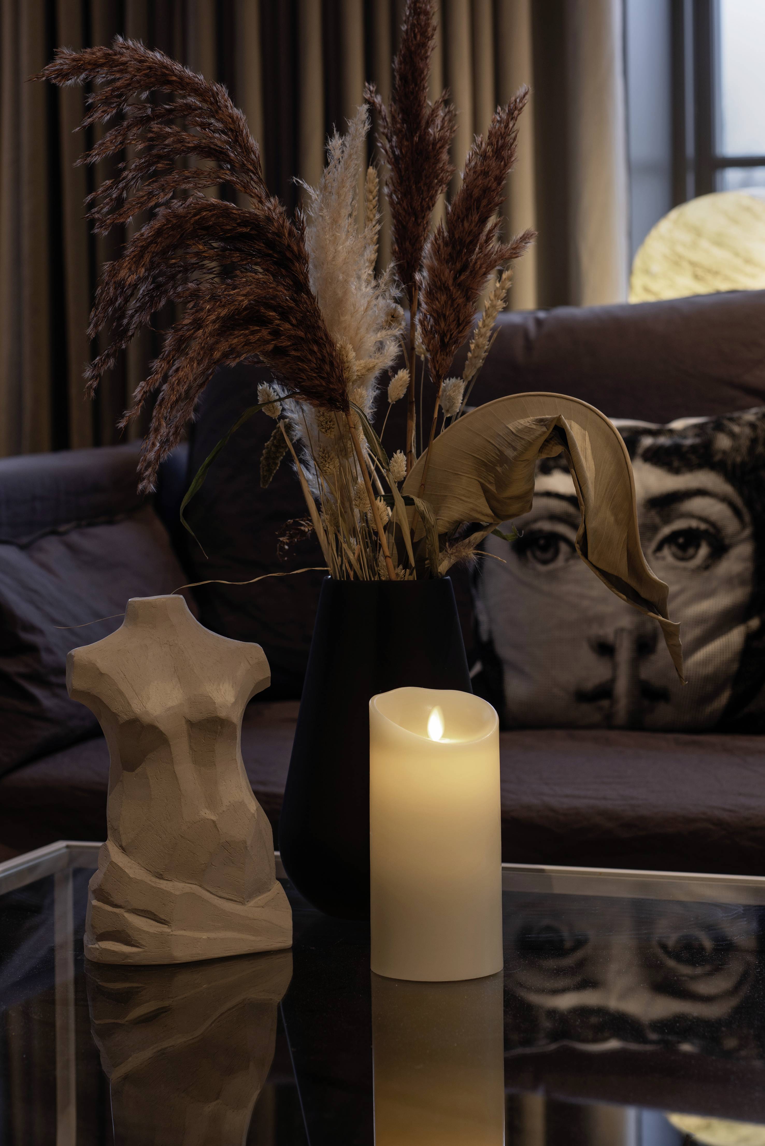 Na stole stoją bezpłomienna świeca, abstrakcyjna rzeźba i wazon z suszonymi trawami przed kanapą.
