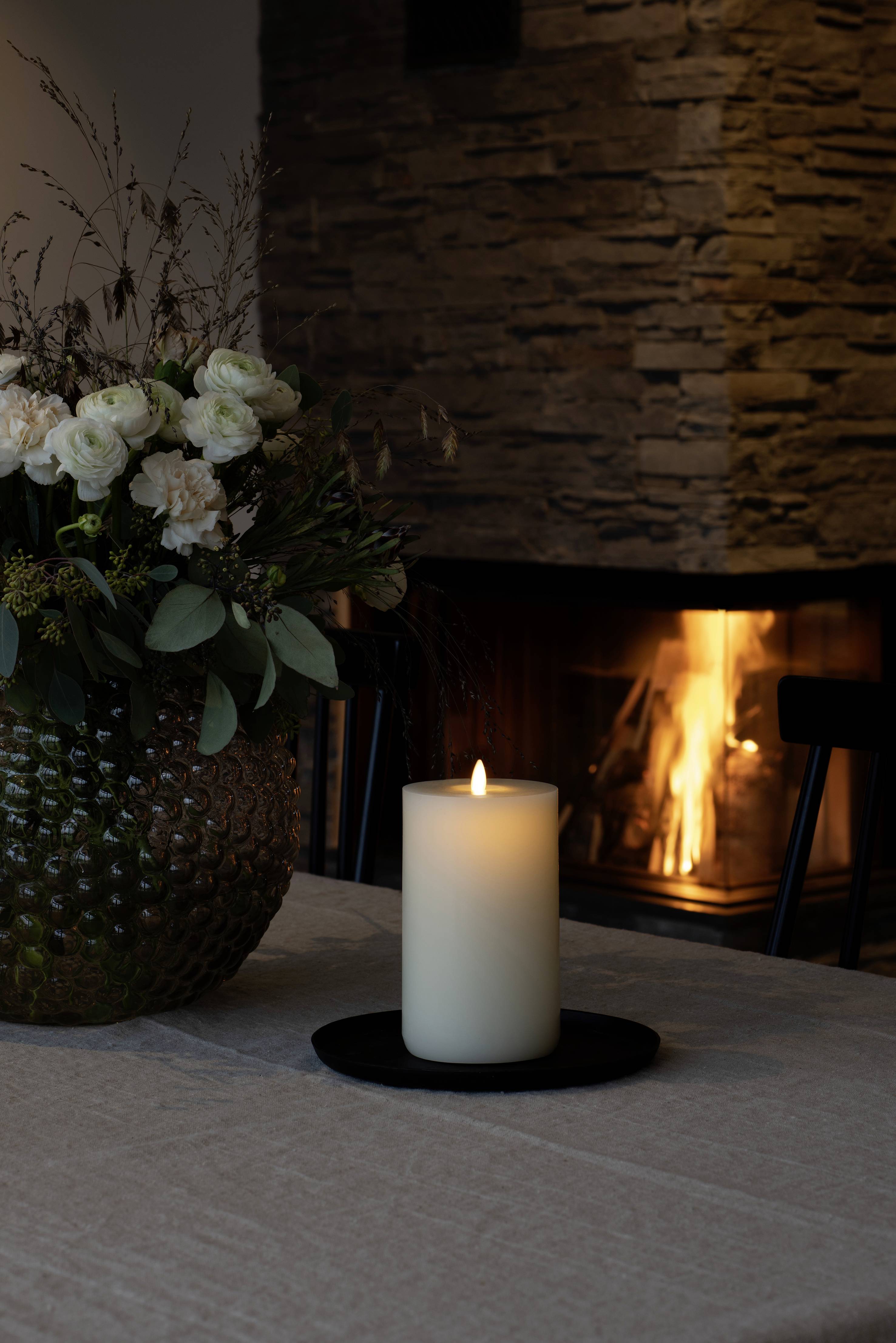 Płonąca świeca stoi na stole przed kominkiem. Obok niej znajduje się wazon ze świeżymi kwiatami.