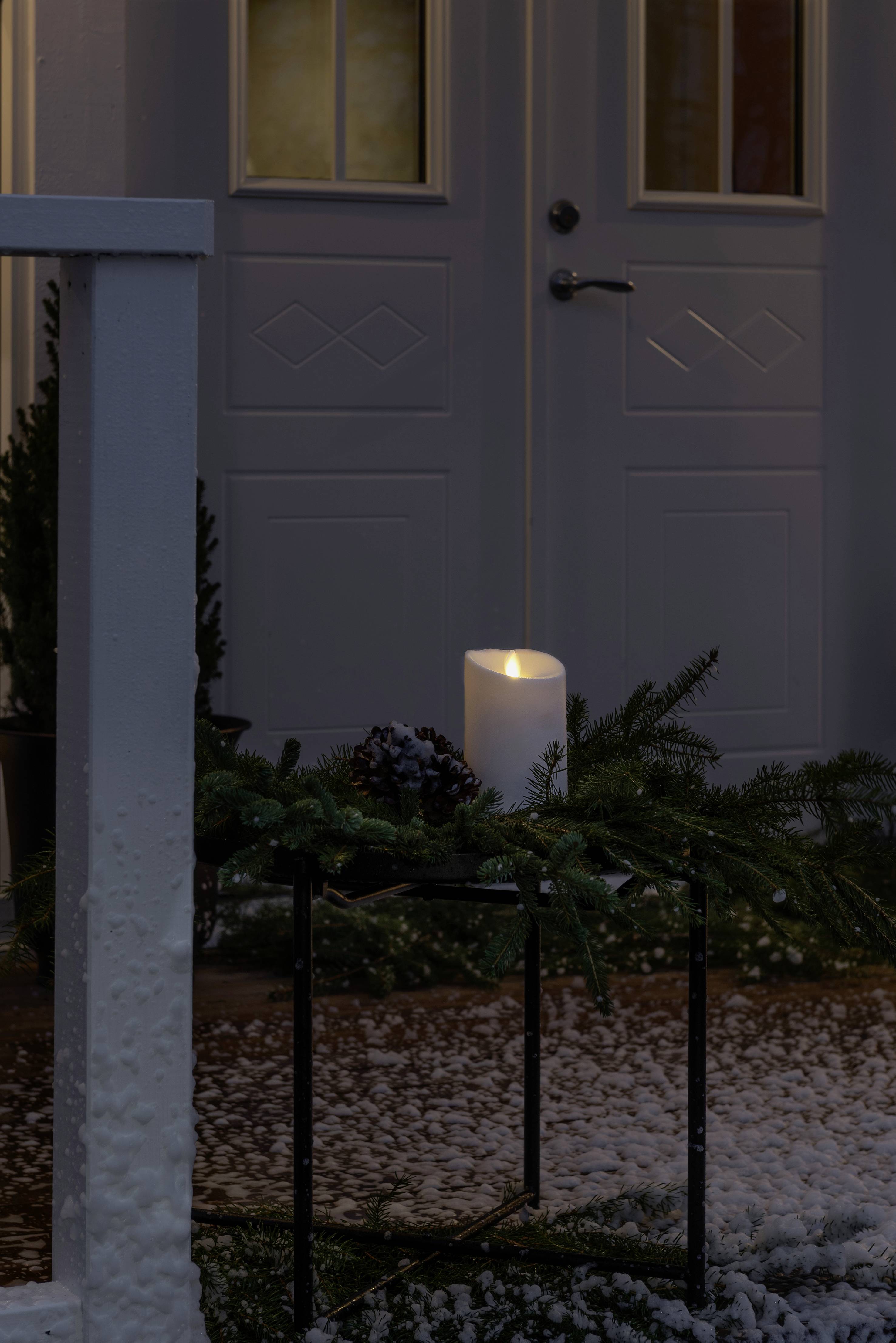 Migocząca biała świeca stoi na stolikach przed drzwiami wejściowymi, otoczona gałązkami sosny i szyszkami, podczas lekkiego opadu śniegu.